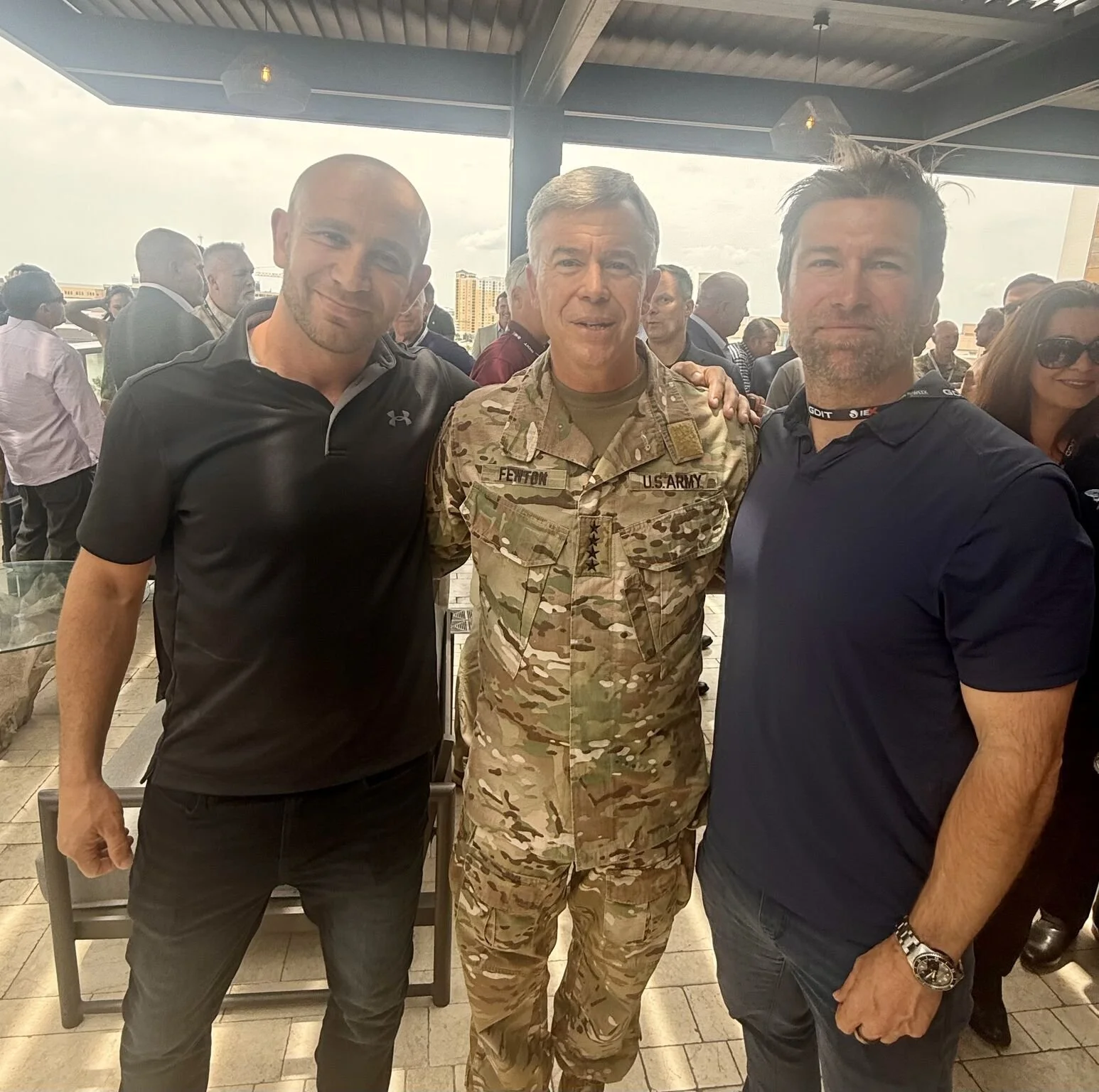 Three men standing together at a social event, with one in camouflage military uniform and the others in casual attire, smiling at the camera.
