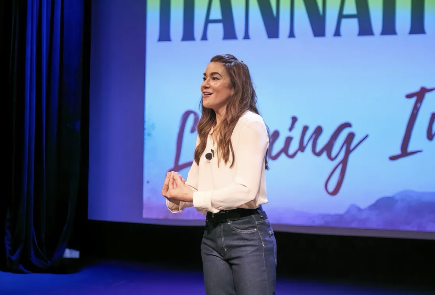 A woman with long wavy hair wearing a white blouse and dark jeans standing on stage, speaking in front of a large screen displaying the words 'Hanna Listening To'.
