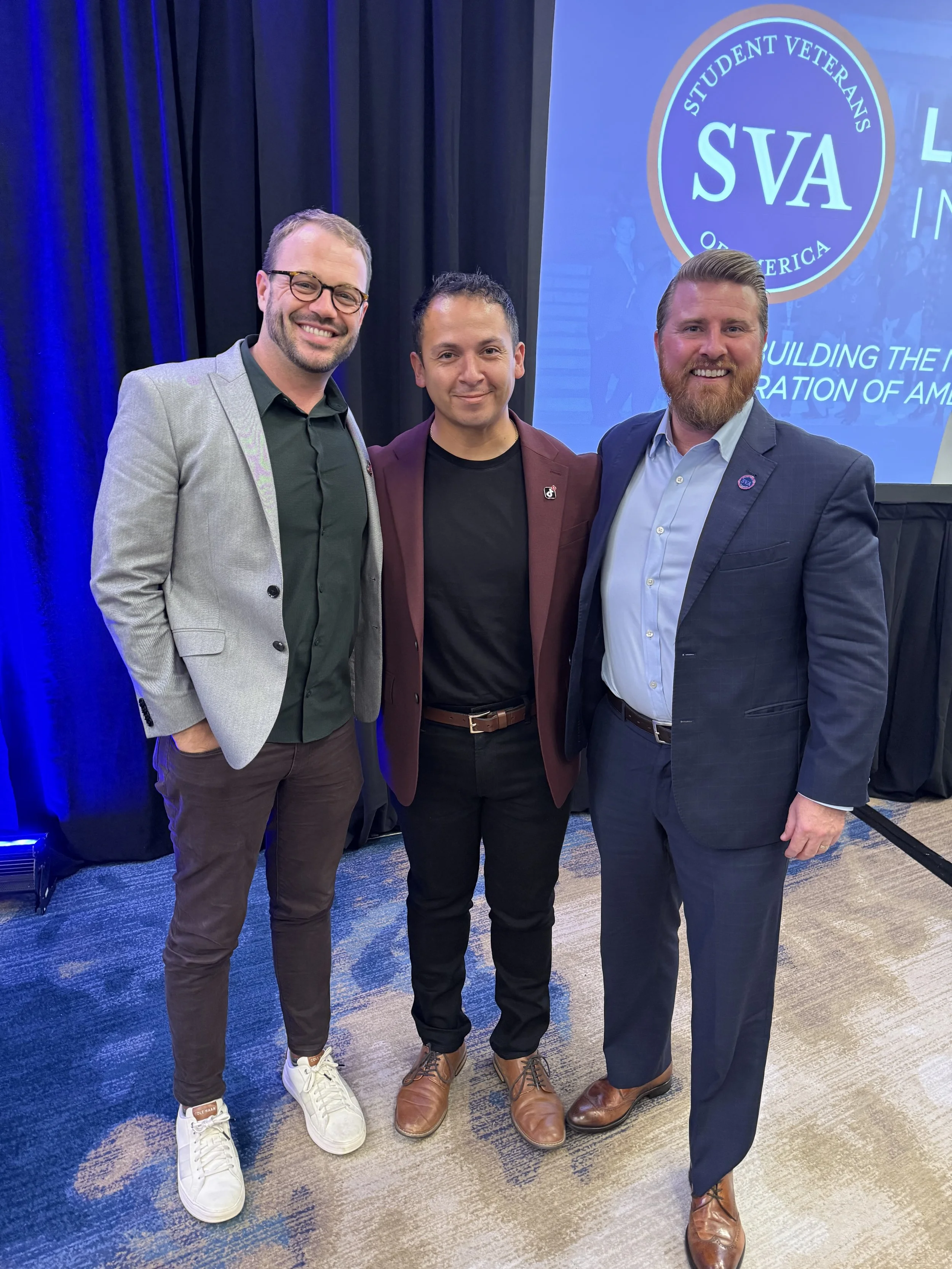 Three men standing together and smiling at an event, with a screen displaying the Student Veterans of America (SVA) logo in the background.