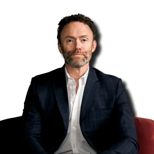 Portrait of a middle-aged man with curly hair and beard, wearing a dark checkered blazer and white shirt, sitting against a black background.