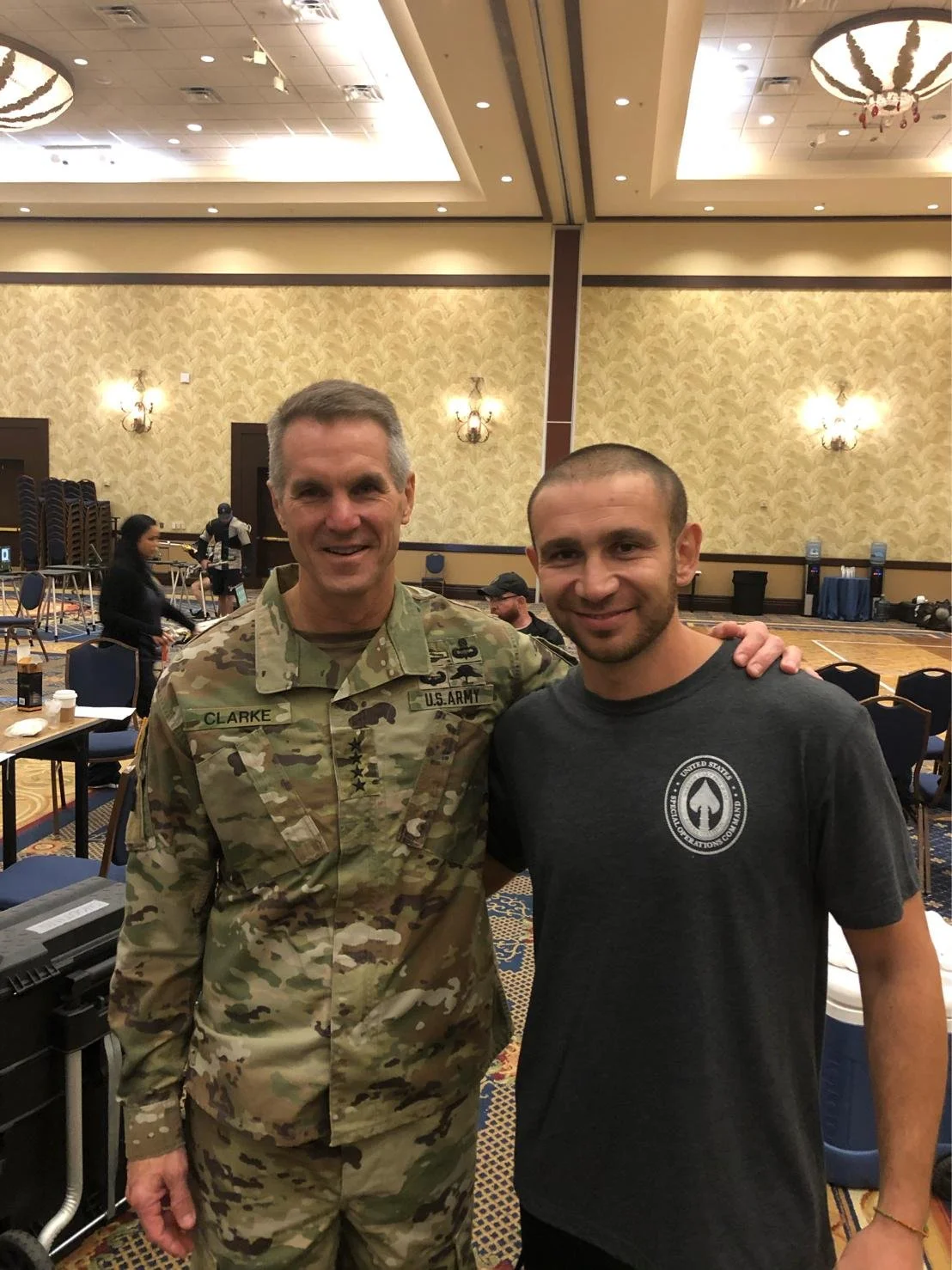 A man in a U.S. Army camouflage uniform standing next to a man in a black t-shirt inside a large, well-lit banquet hall with decorative wallpaper and chandeliers.