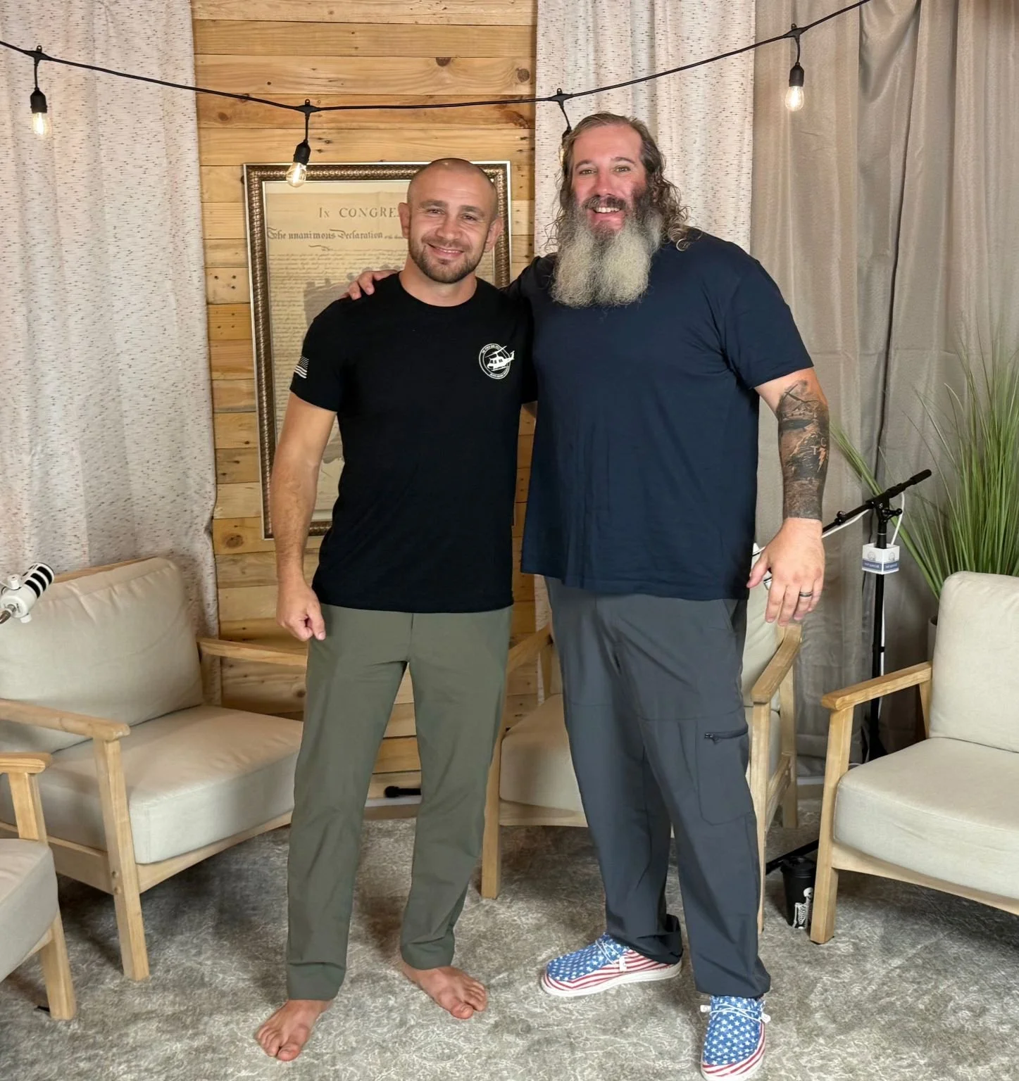 Two men standing together and smiling. one of them has his arm around the other's shoulder. The man on the left has a shaved head, beard, and is barefoot, wearing a black t-shirt and light gray pants. The man on the right has long, curly gray hair an