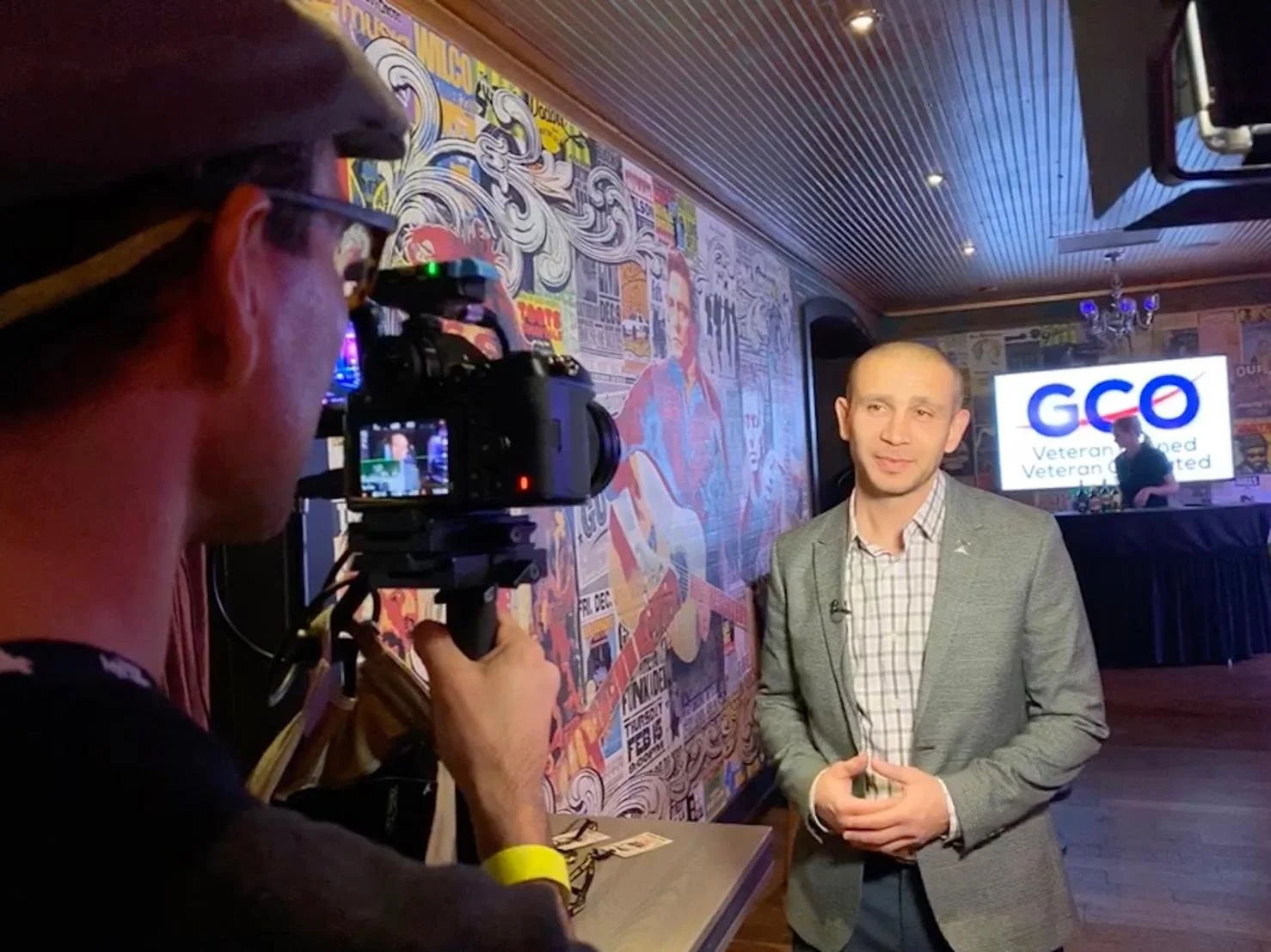 A man in a gray blazer and plaid shirt is being interviewed or giving a speech in front of a television screen that displays the GCO logo and the words "Veteran Owned, Veteran Operated." A photographer is capturing the moment with a camera. The backg