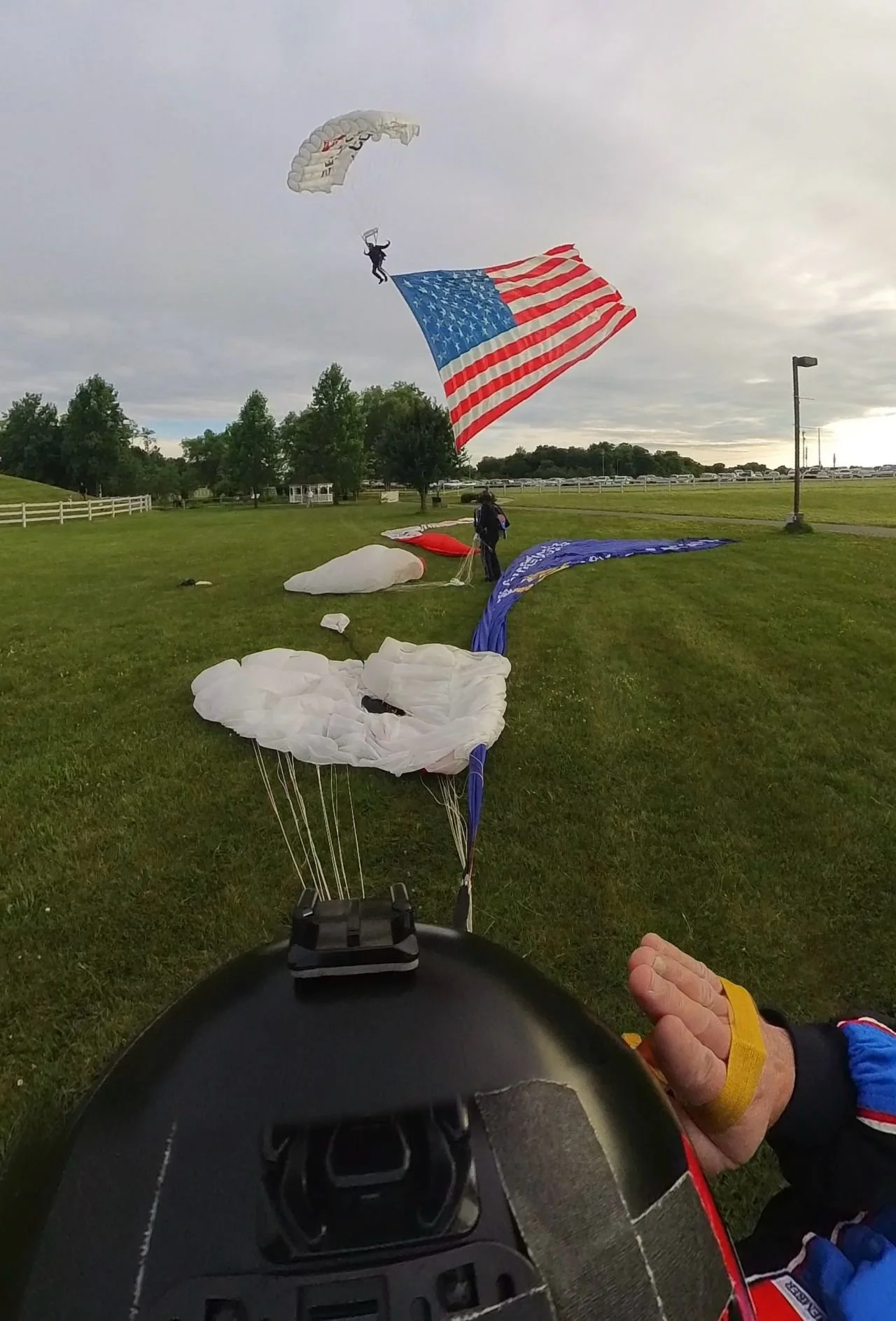 A skydiver with an American flag parachute descending onto a grassy field, with its colors and stars visible, and a person on the ground preparing for landing.