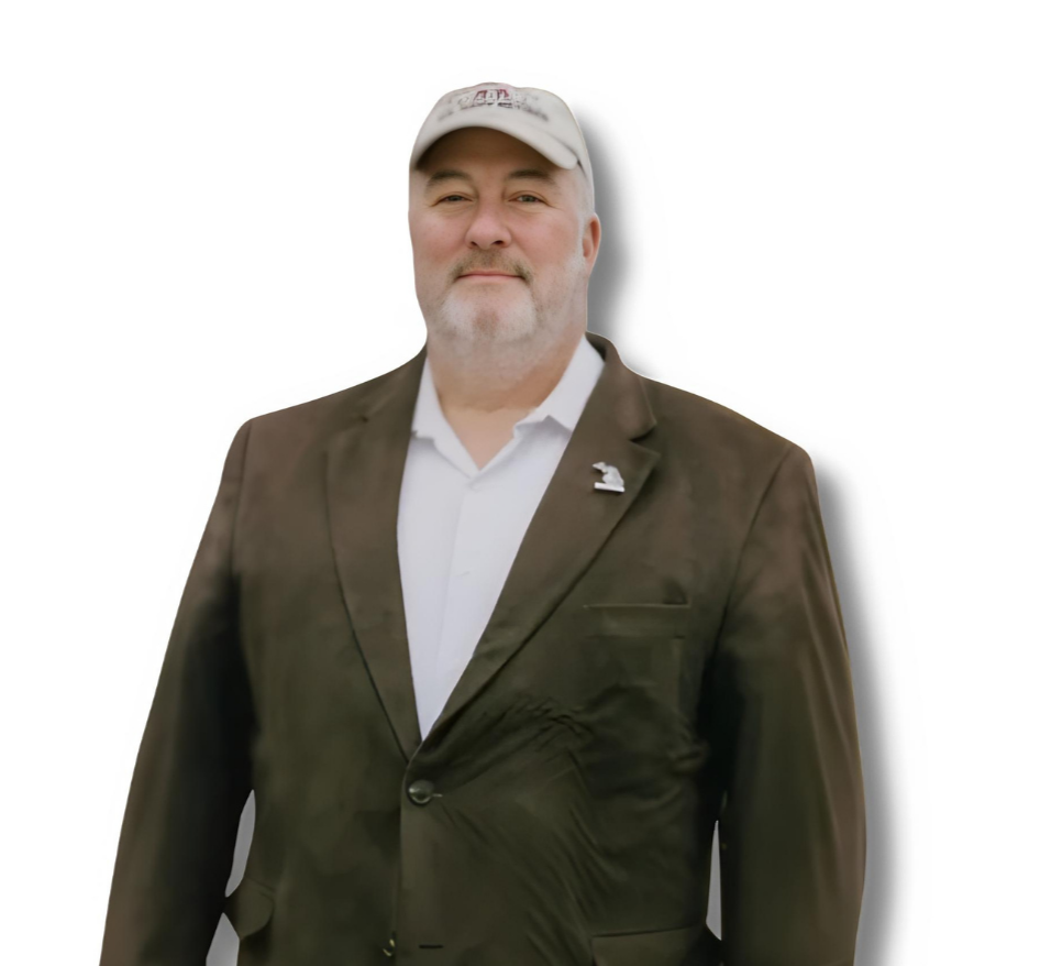 A middle-aged man wearing a white baseball cap, white dress shirt, and dark blazer with a lapel pin, standing against a plain white background.