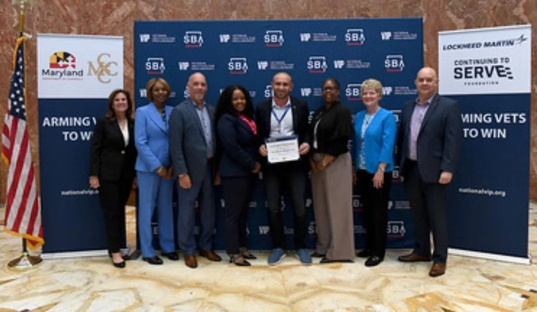 Group of seven diverse people standing together at an event, with banners and an American flag in the background. One person is holding a certificate.