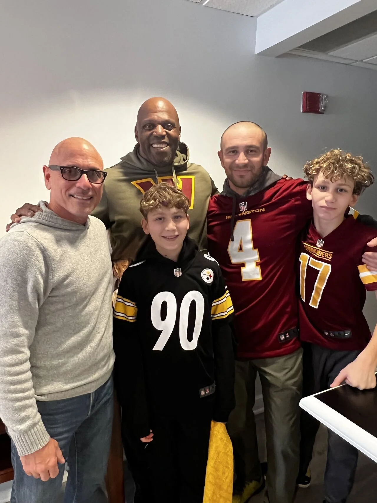 A group of five people, including two children, posing for a photo indoors. Two of them are wearing football jerseys from the Washington Football Team, and the other two adults are dressed casually. Everyone is smiling.