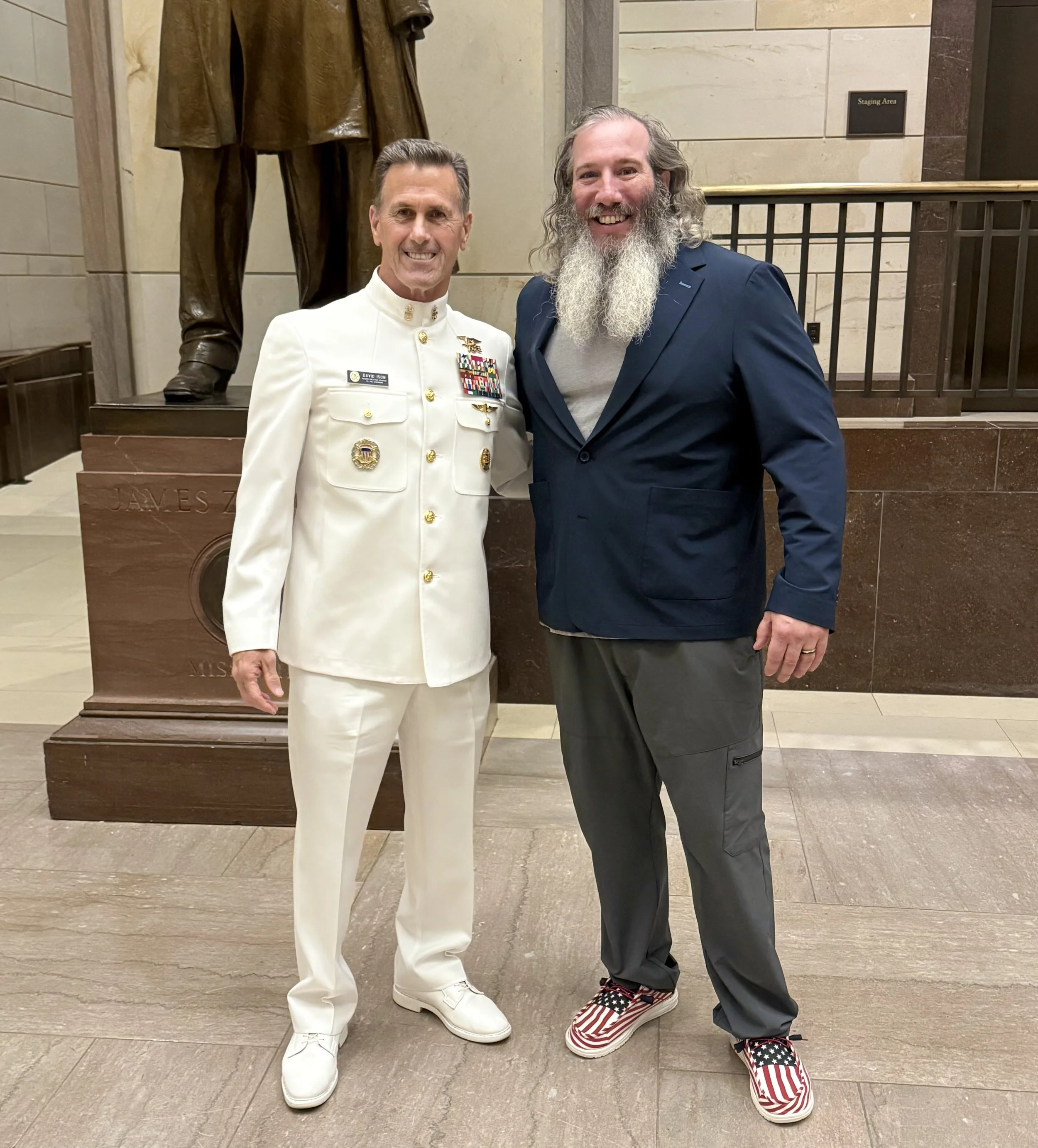 Two men standing together in a public building, smiling. The man on the left is wearing a white military uniform with medals and gold buttons, while the man on the right is dressed casually in a blue blazer, gray T-shirt, gray pants, and patriotic sn