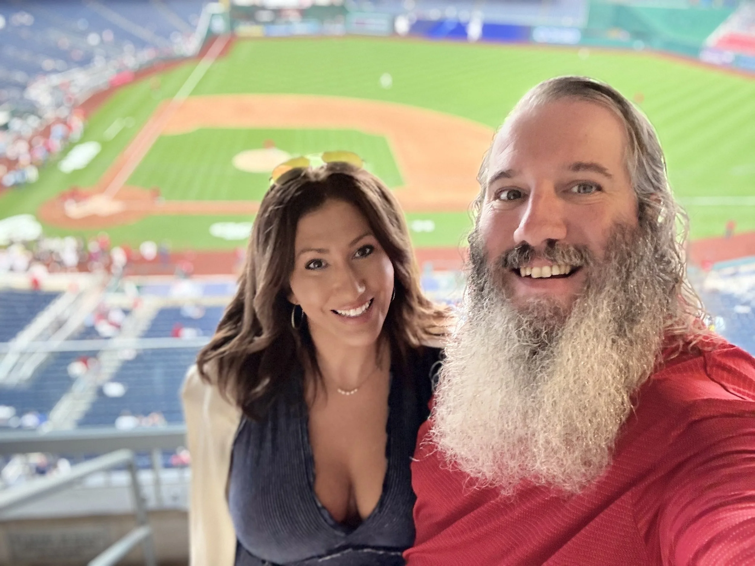 A man with a gray beard and long hair smiling in front of a stadium baseball field, with a woman with long brown hair smiling beside him.