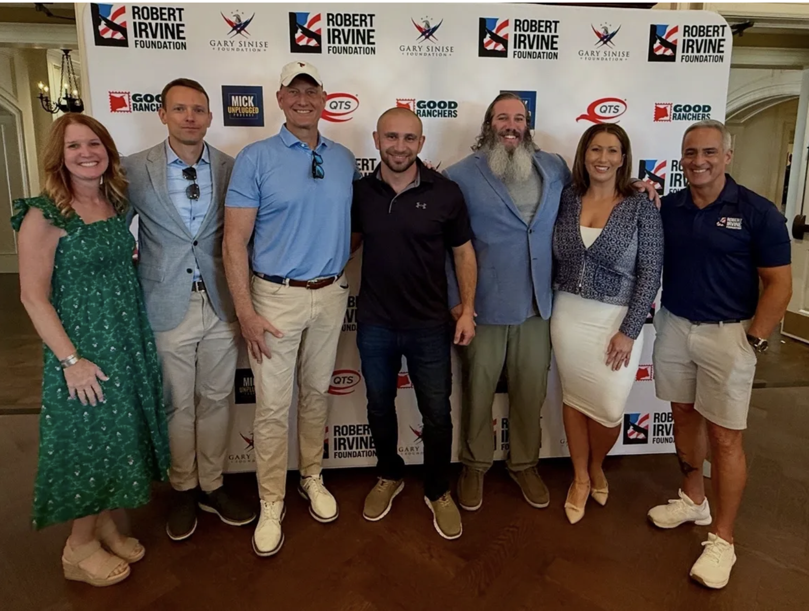 Group of eight people standing in front of a backdrop with logos, smiling and posing for a photo. The backdrop features logos of the Robert Irvine Foundation, Gary Sinise Foundation, Good Ranchers, QTS, and Mick Unplugged.