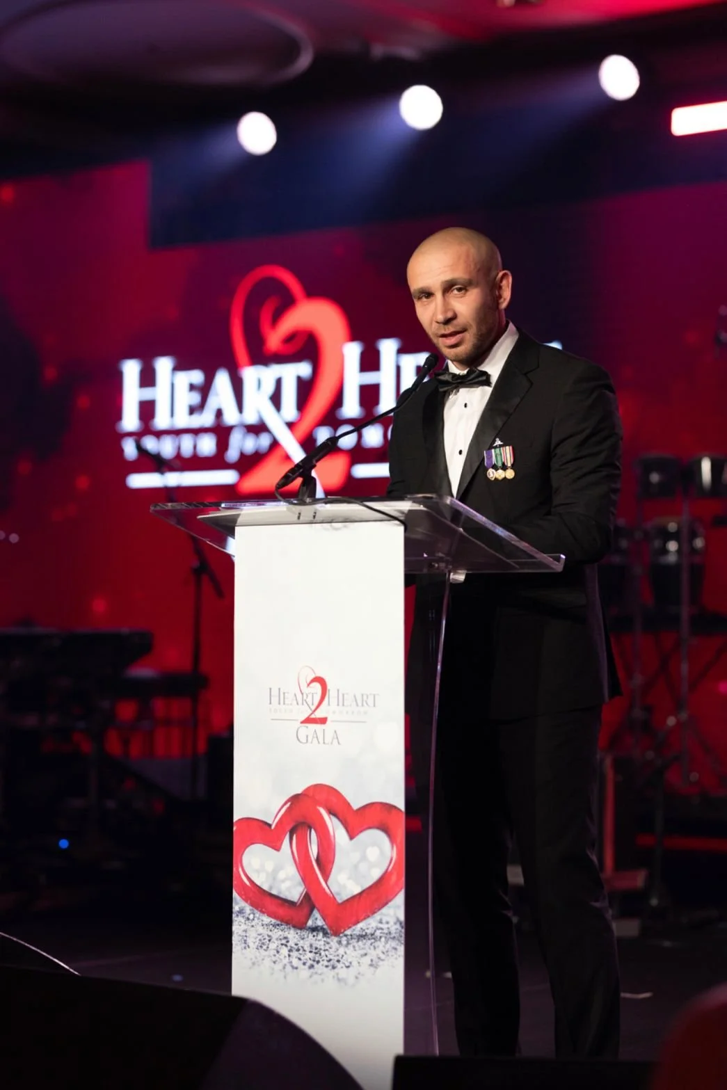 Man in tuxedo with medals speaking at a podium during a charity event, with a background displaying a red heart logo and the text "Heart 2 Heart" and "Gala."
