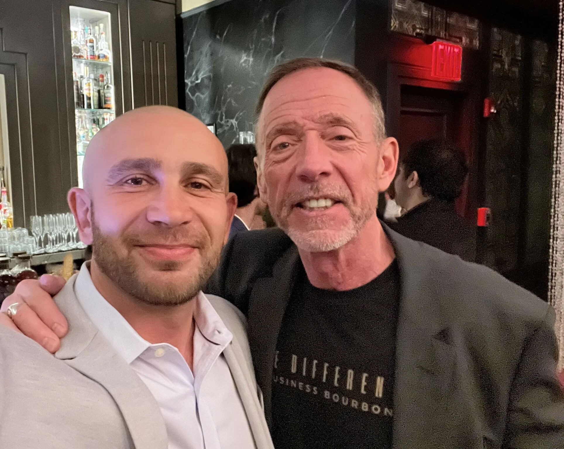 Two smiling men taking a selfie together in a dimly lit bar or restaurant. One man is bald and wearing a light-colored shirt, and the other has gray hair and a beard, and is wearing a darker shirt. There are liquor bottles visible in the background a
