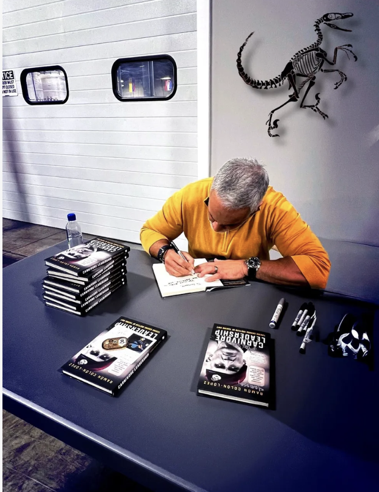 Author sitting at table signing copies of his book, with a stack of books, markers, and a water bottle nearby. A dinosaur skeleton drawing is on the wall behind him.