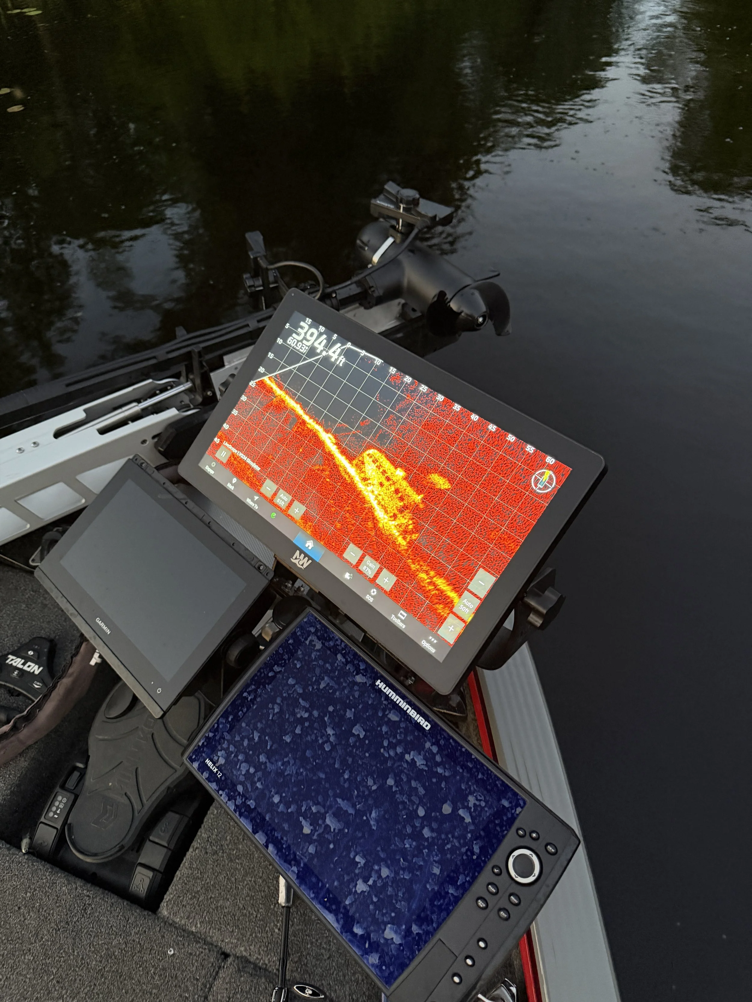 Multiple electronic devices and monitors displaying sonar readings and data, set up on a boat's dashboard near a body of water with reflections of trees.