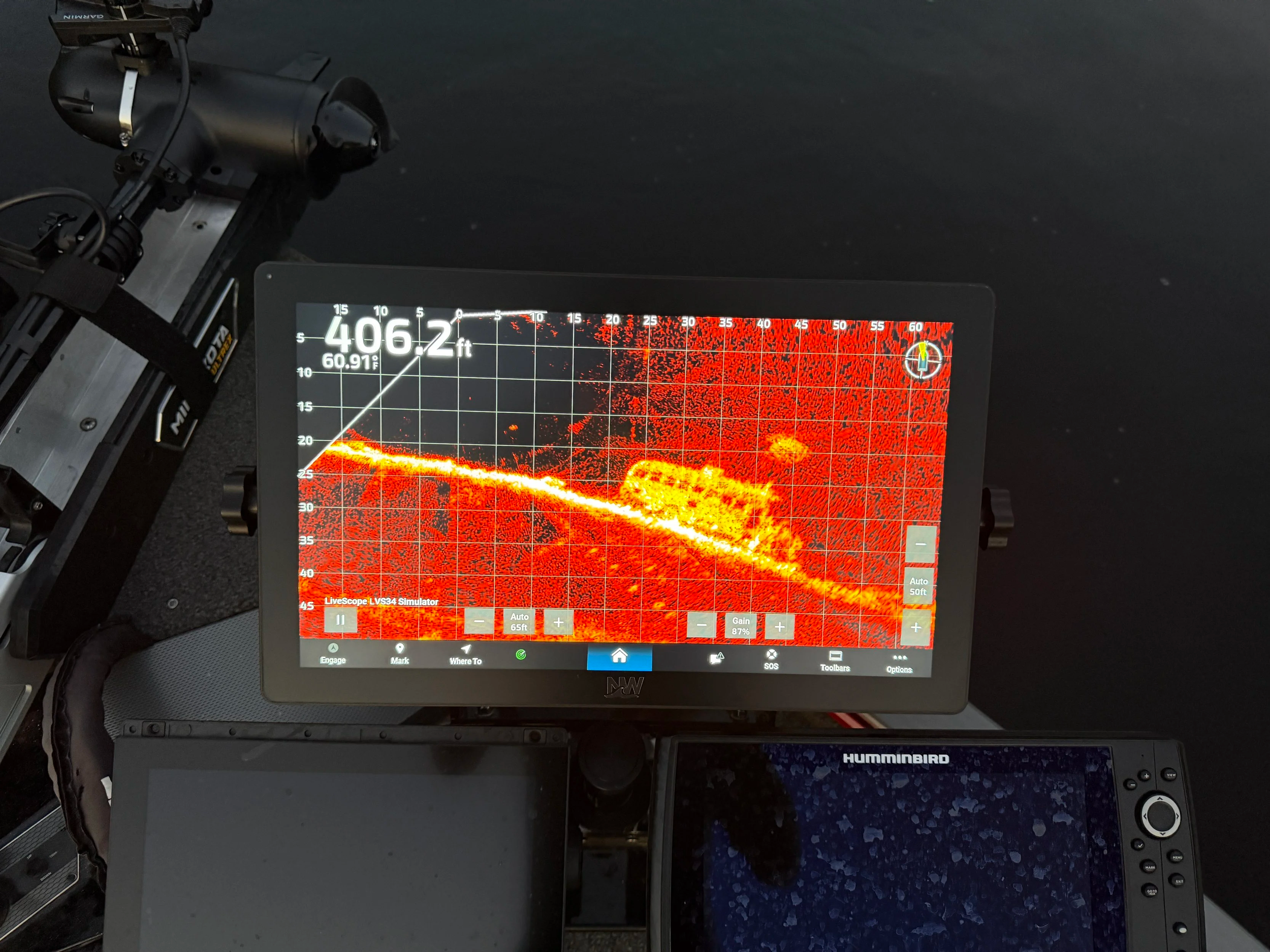 A sonar or fishfinder screen displays a curved, bright yellow and orange object with surrounding red dots on a black background, indicating underwater structures or fish. The device is connected to a metal mounting hardware on the left and is part of a boat or marine setup.