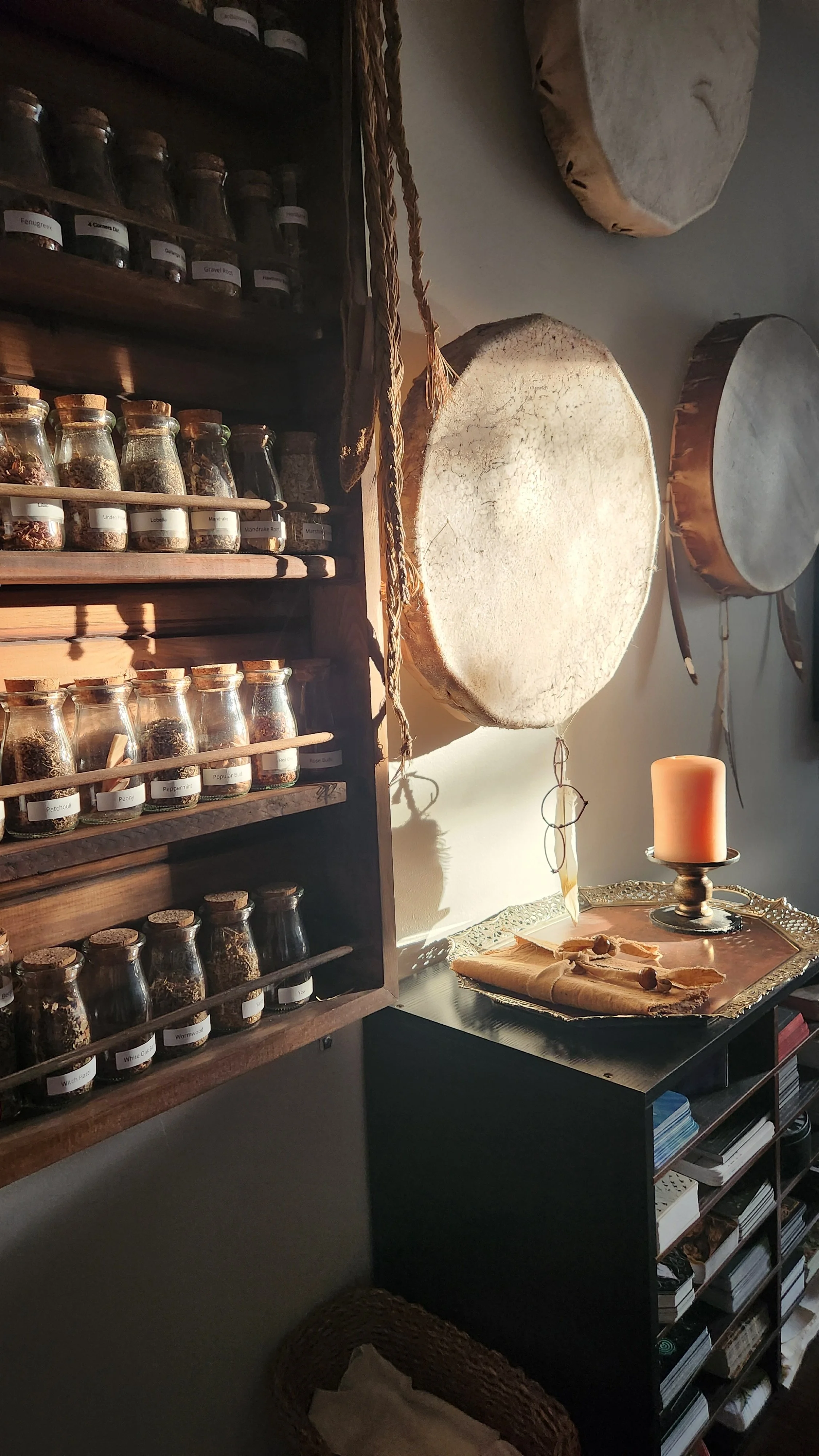 A wooden spice rack with small glass jars labeled with various herbs and spices, a round table with a gold tray holding rolled paper and objects, a peach-colored candle in a holder, a large circular drum on the wall, and a basket on the floor.