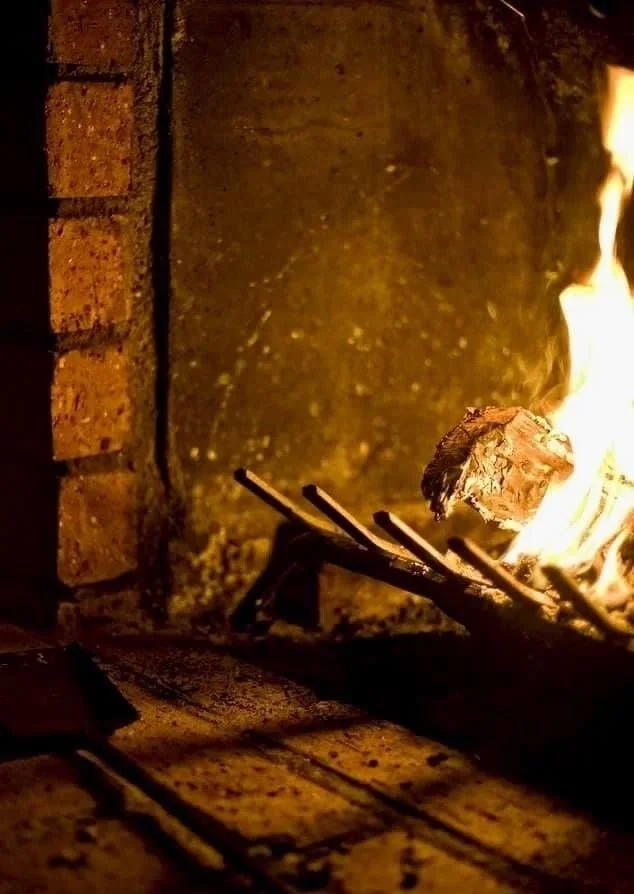 A close-up of a brick fireplace, a hearth with a small fire burning on a grate, illuminating the bricks and surrounding area.
