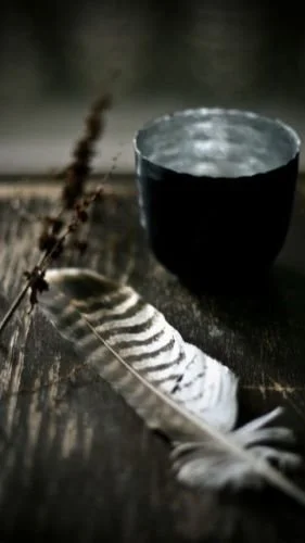 A black cup of water, a feather, and dried herbs on a wooden surface.
