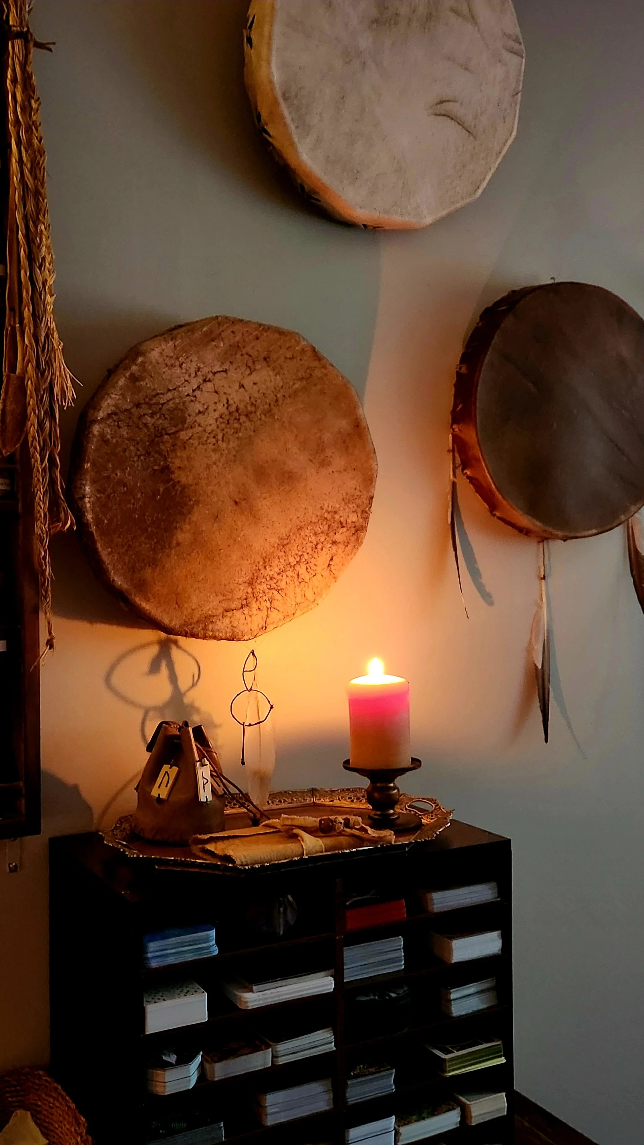 A wall decorated with three large round drums, a lit candle on a black table with decorative objects, and a cabinet filled with various books and stacks of papers.