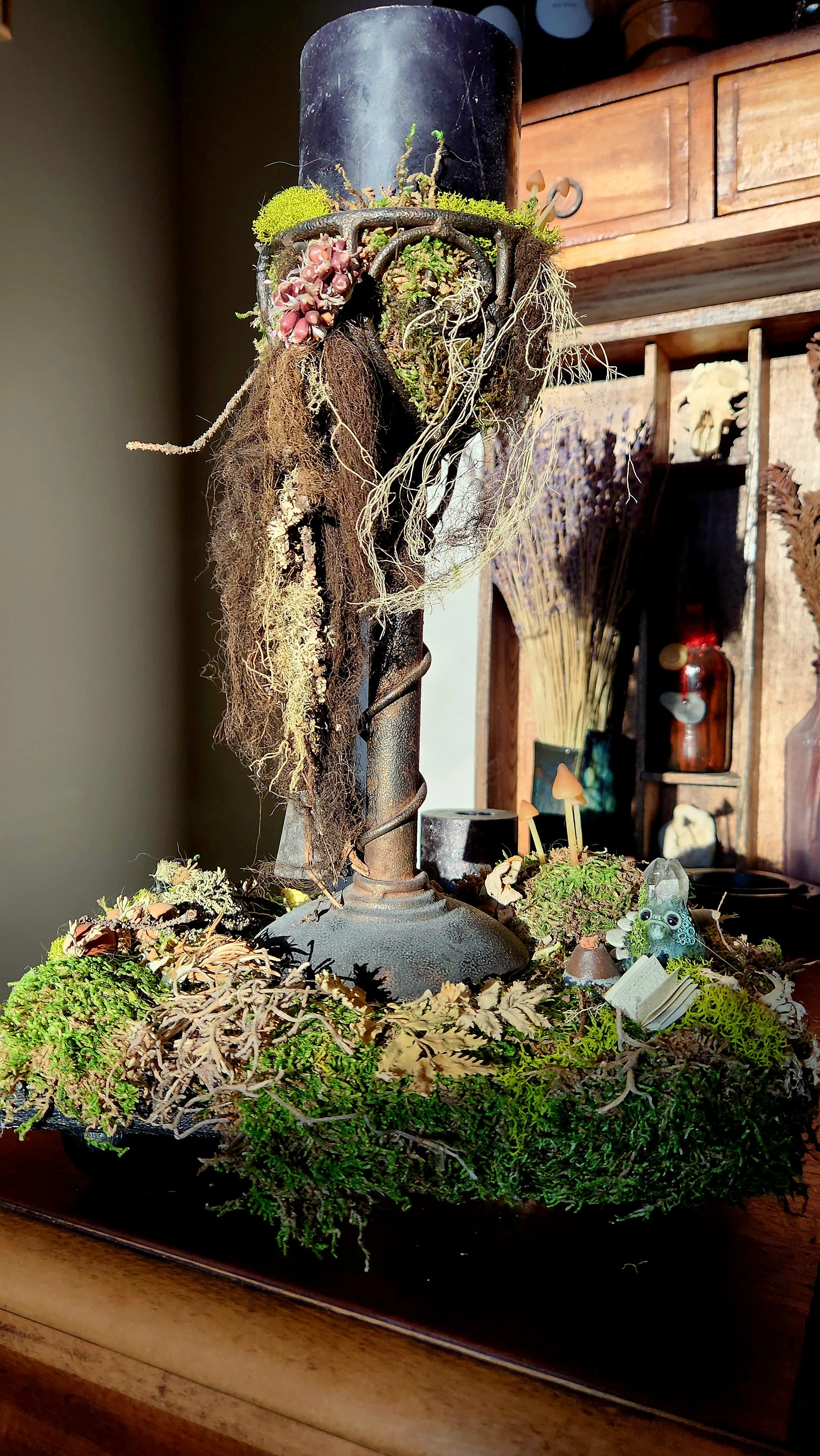 A decorative arrangement featuring moss, dried plants, and small mushrooms on a metal stand, with a wooden shelf and various ornaments in the background.