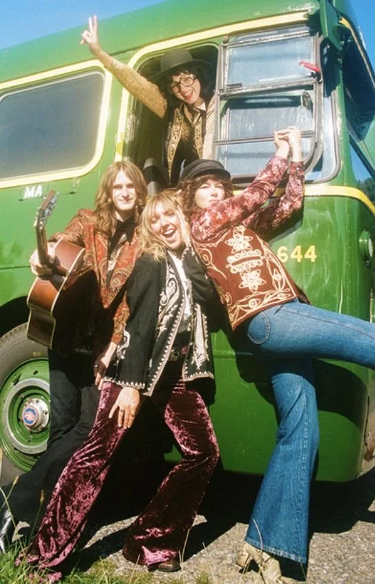 3 women and one man with long hair dressed in vintage clothing posing in front of a green and yellow bus as one woman peeks out of the window.