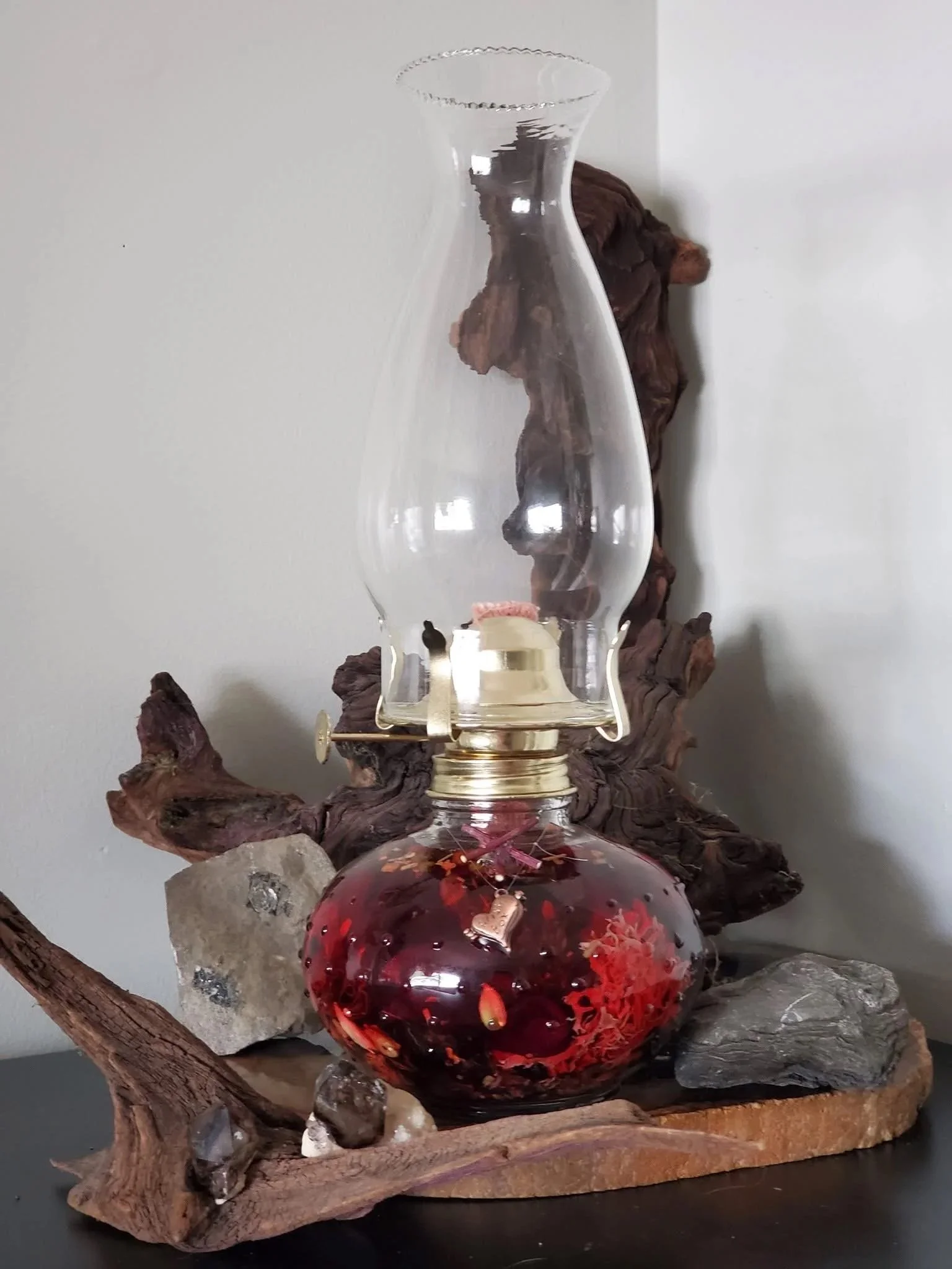 A magic glass oil lamp with a metal burner, sitting on a driftwood base, with red wax candle inside the glass bottom, surrounded by rocks.