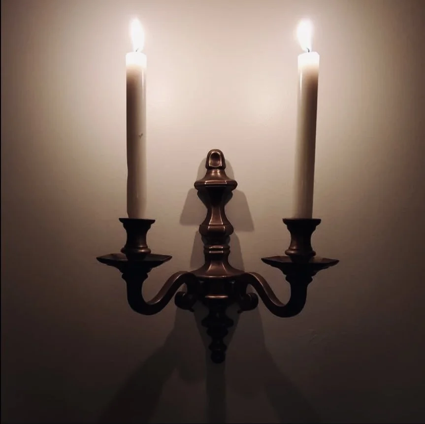 A bronze wall-mounted candle sconce with two lit candles illuminating a plain wall.