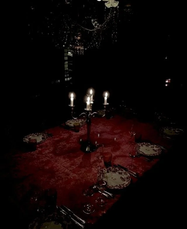 A dining table set with a red tablecloth, surrounded by plates, glasses, and silverware. A chandelier hangs above, and a candelabrum with lit candles is centered on the table, creating a dimly lit, elegant ambiance.