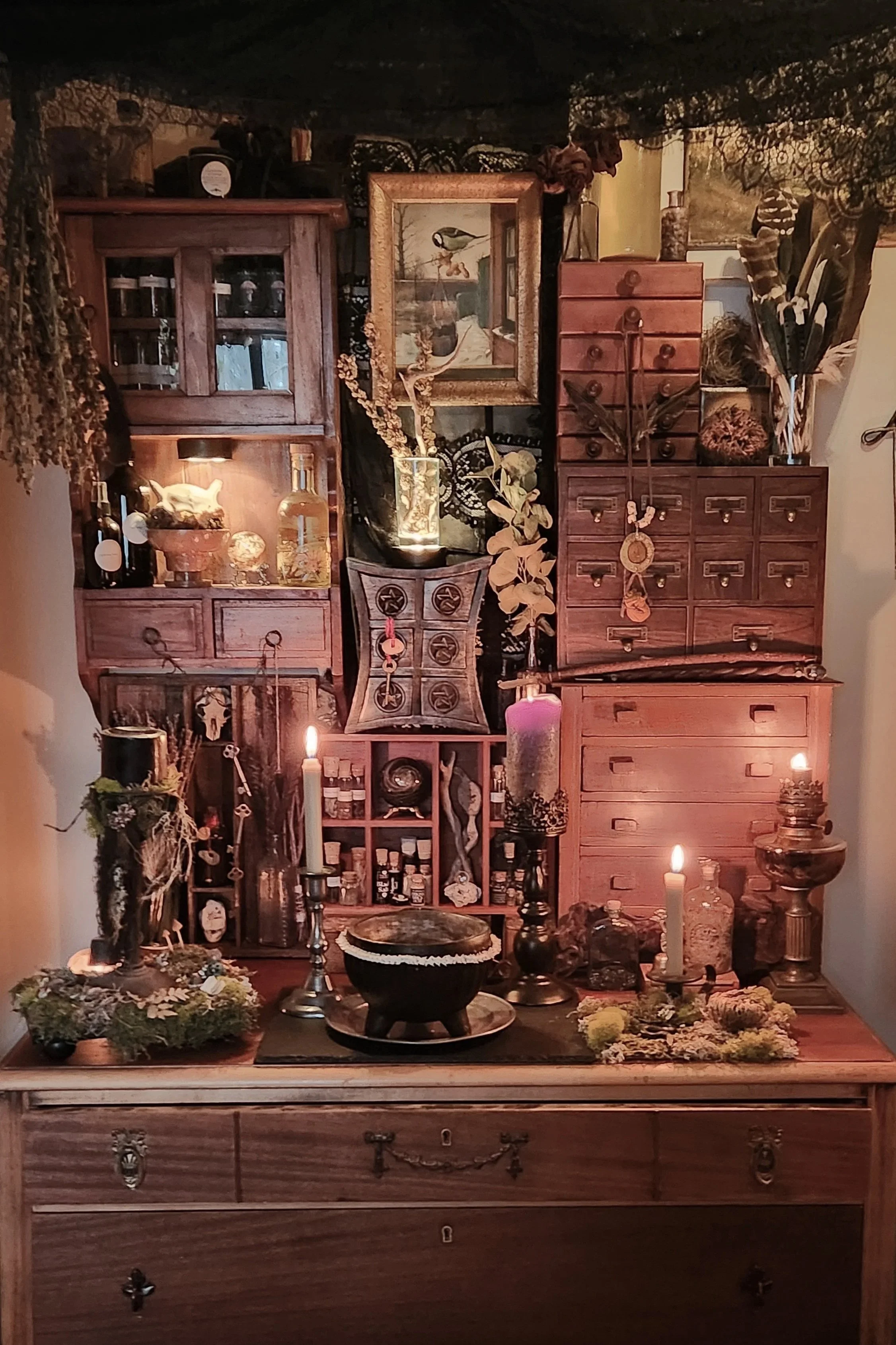 A wooden display cabinet filled with various decorative items, including plants, candles, and art pieces, set against a black lace backdrop with additional decor and candles on the table in front.