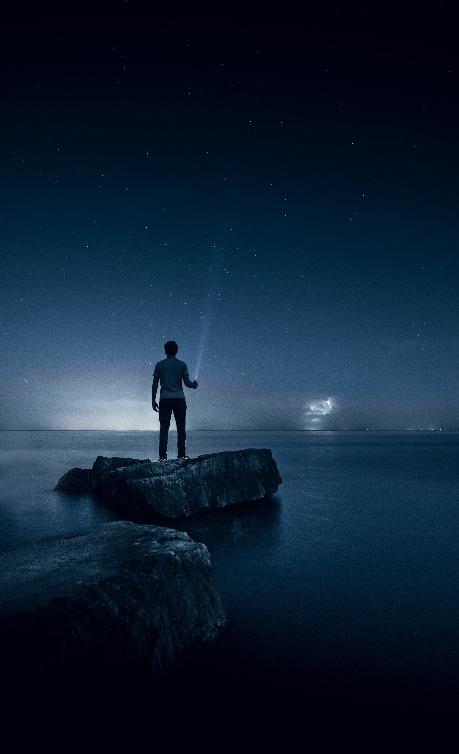 A person standing on a large rock by the ocean at night, holding a flashlight and gazing at the starry sky.