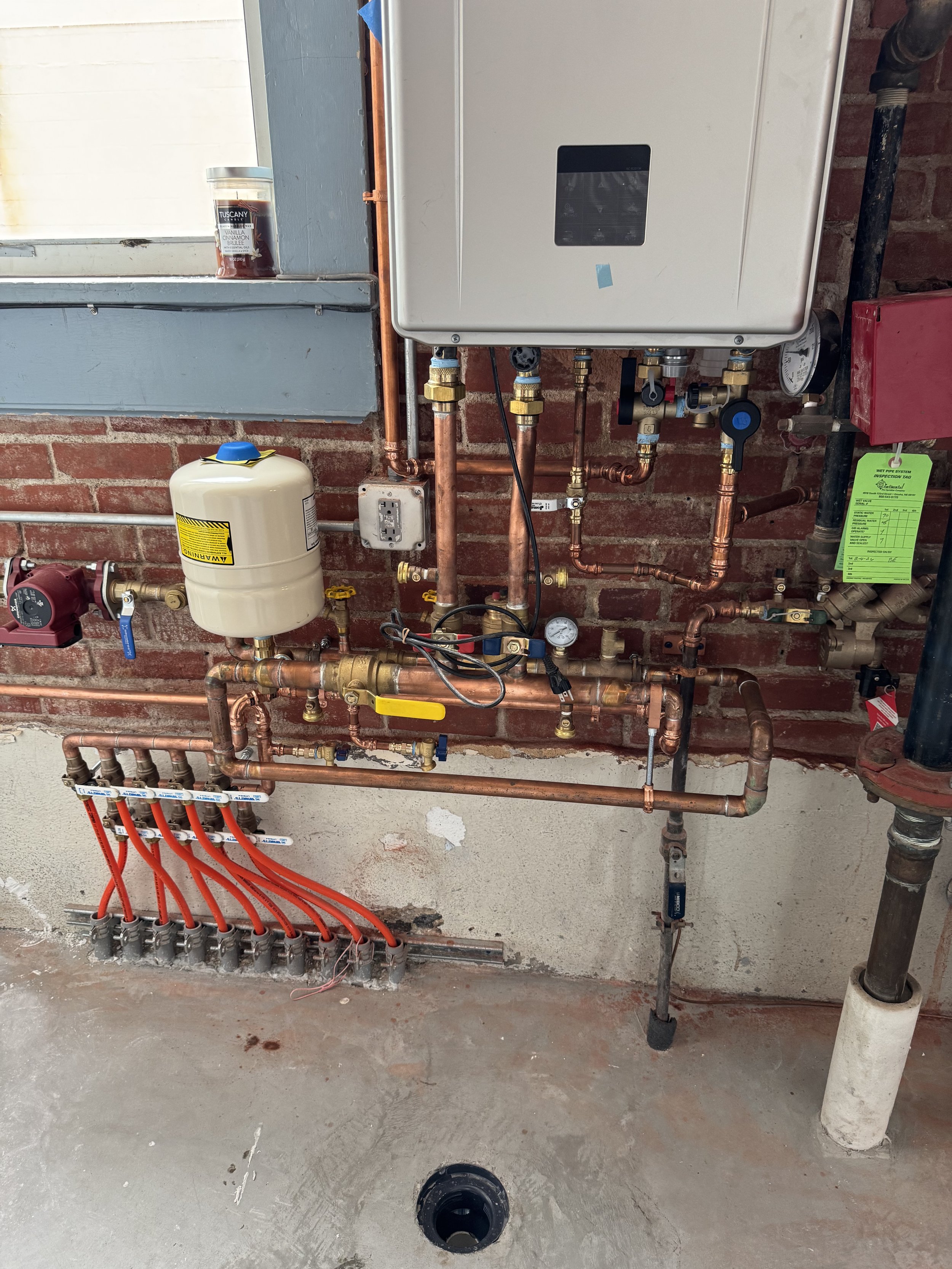 A plumbing setup on a brick wall with copper pipes, various valves, a white water filtration tank with red pipes connected at the bottom, and an electrical panel with a digital display.