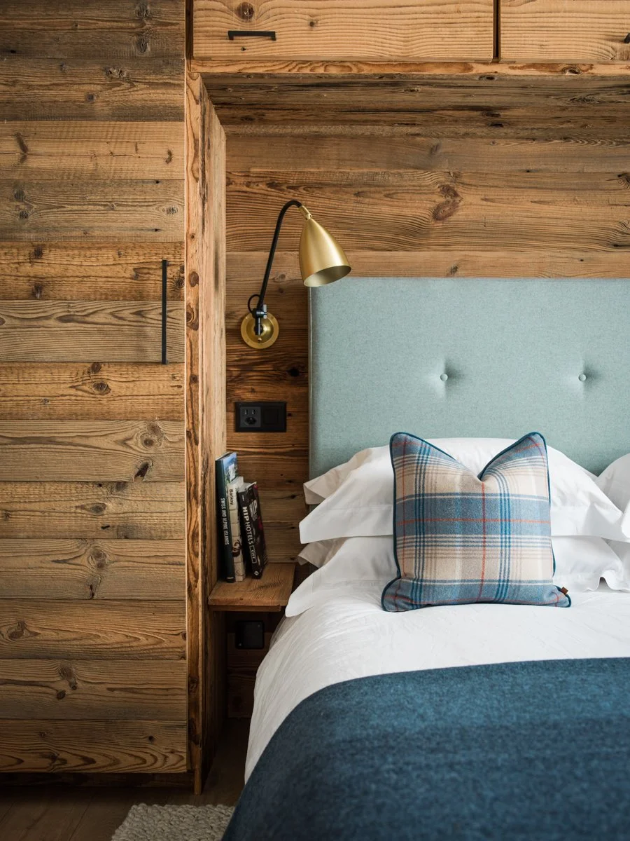 Close-up of a cozy bedroom with a wood-paneled wall, a bed with white linens, a plaid throw pillow, a light blue upholstered headboard, a wall-mounted gold reading lamp, and a small bedside shelf with books.