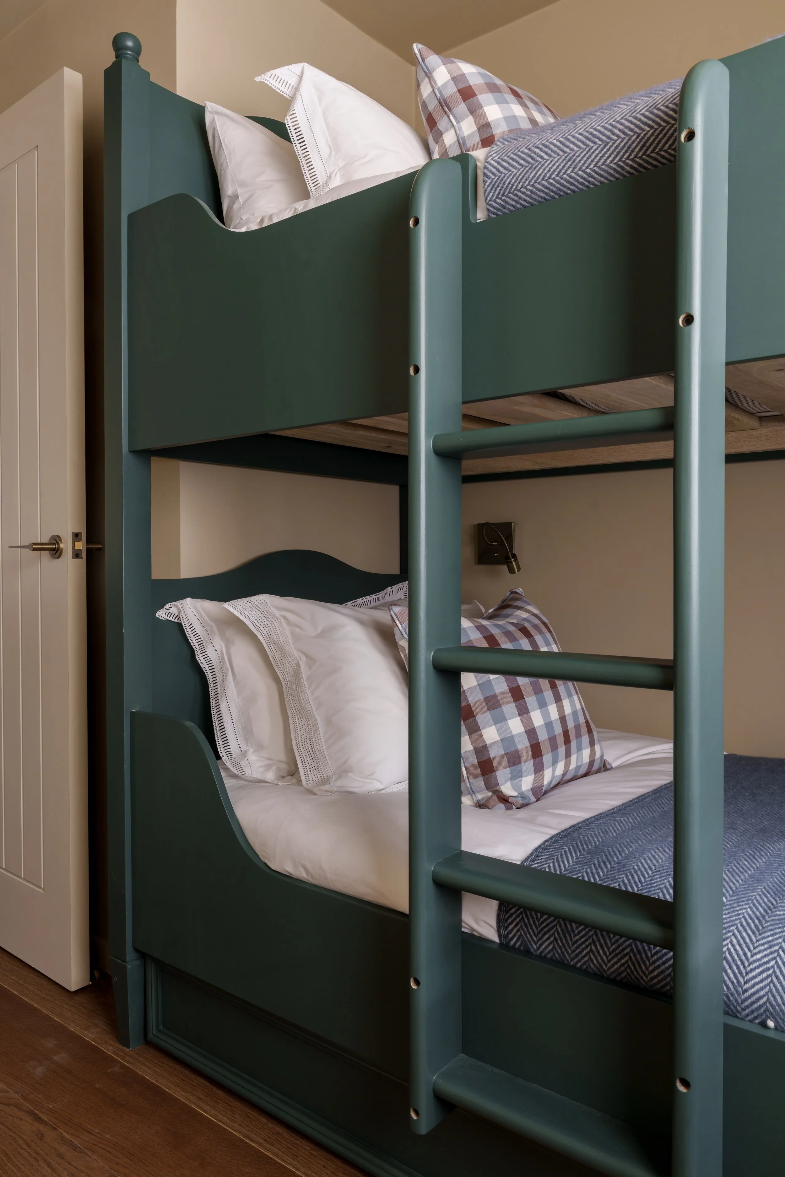 A top bunk bed with white bedding and pillows, a plaid pillow on the lower bunk, and a ladder on the side, next to a closed beige door.