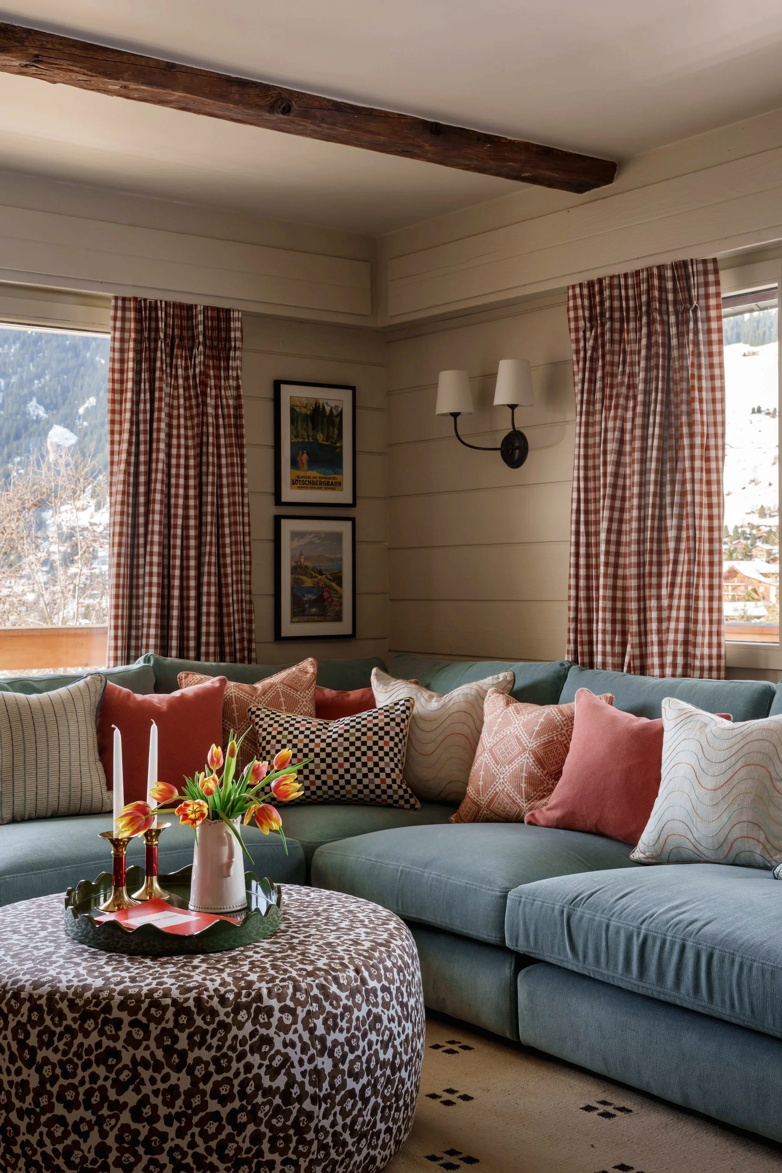 Cozy living room with a teal sectional sofa adorned with various patterned and solid-colored pillows. A leopard print ottoman serves as a coffee table with a decorative tray, a vase with tulips, and candlesticks. The room features light wood-paneled 