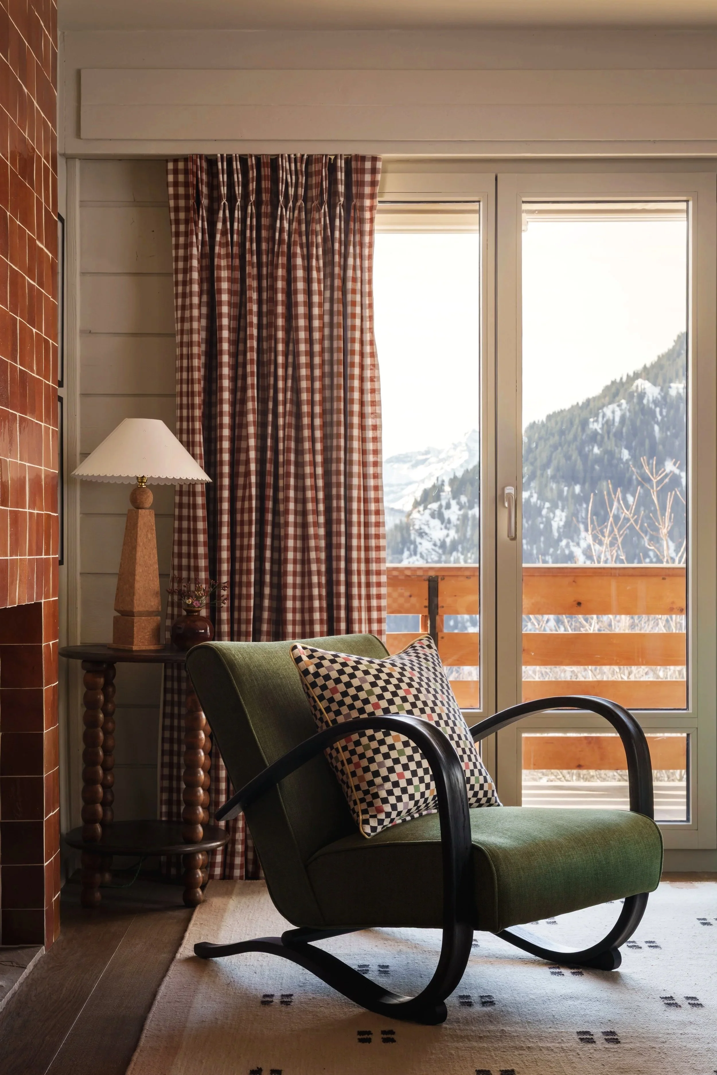 Cozy living room corner with a green rocking chair and checkered pillow, next to a wooden side table with a lamp, overlooking snowy mountain landscape through large glass doors with red checkered curtains.