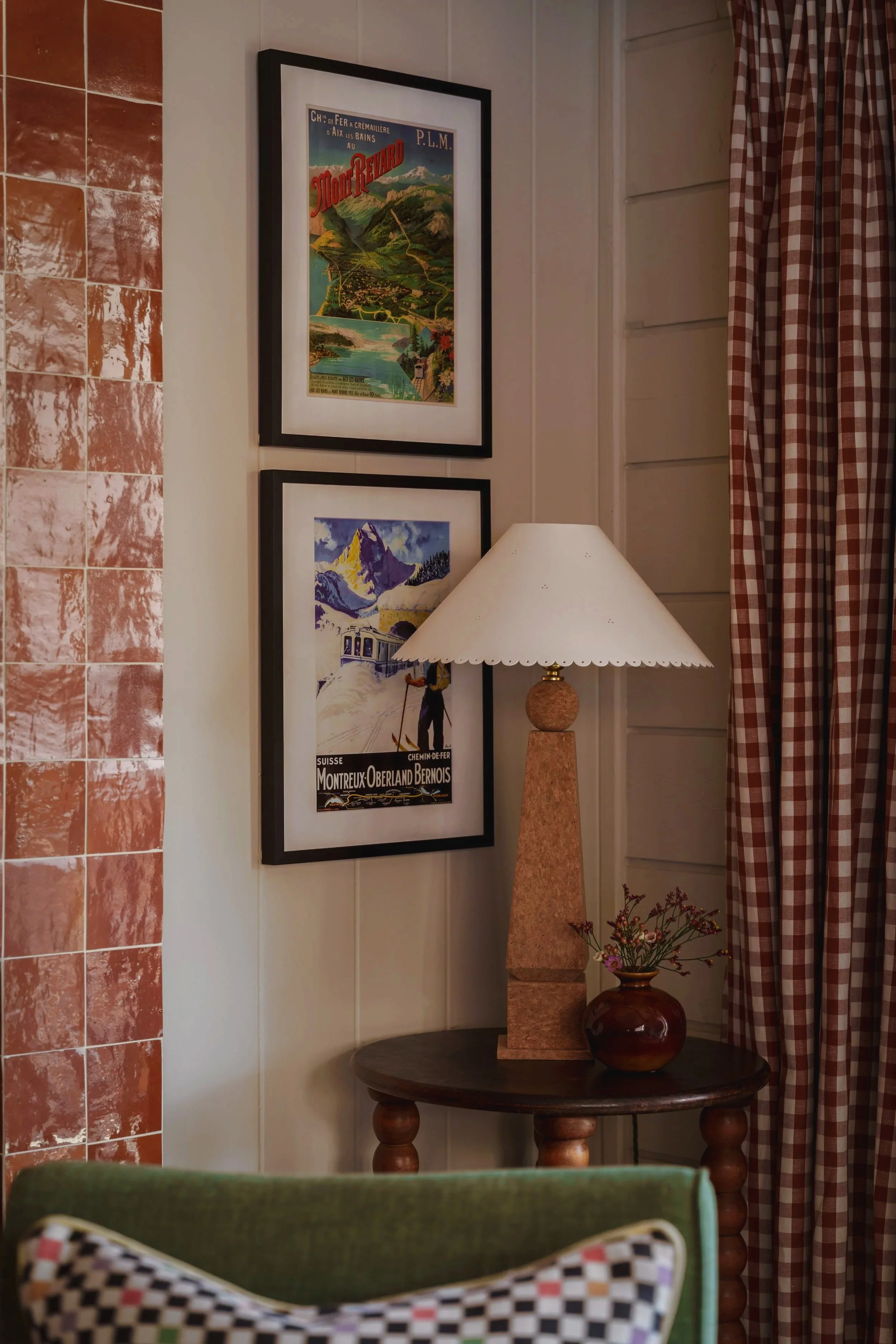 Corner of a room showing two framed vintage travel posters on a white wall, a table with a decorative lamp, a small vase with flowers, and a checkered curtain.