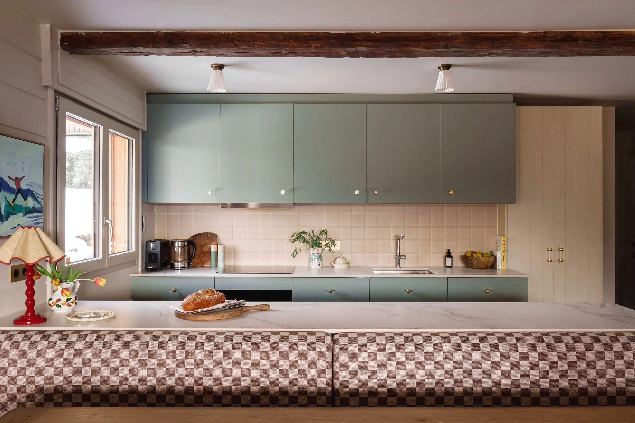 Cozy kitchen with pastel green cabinets, a marble countertop, and a checkered brown and beige bench in front. Decor includes a red table lamp, vase with tulips, bread on a wooden board, and various small items on the counter.
