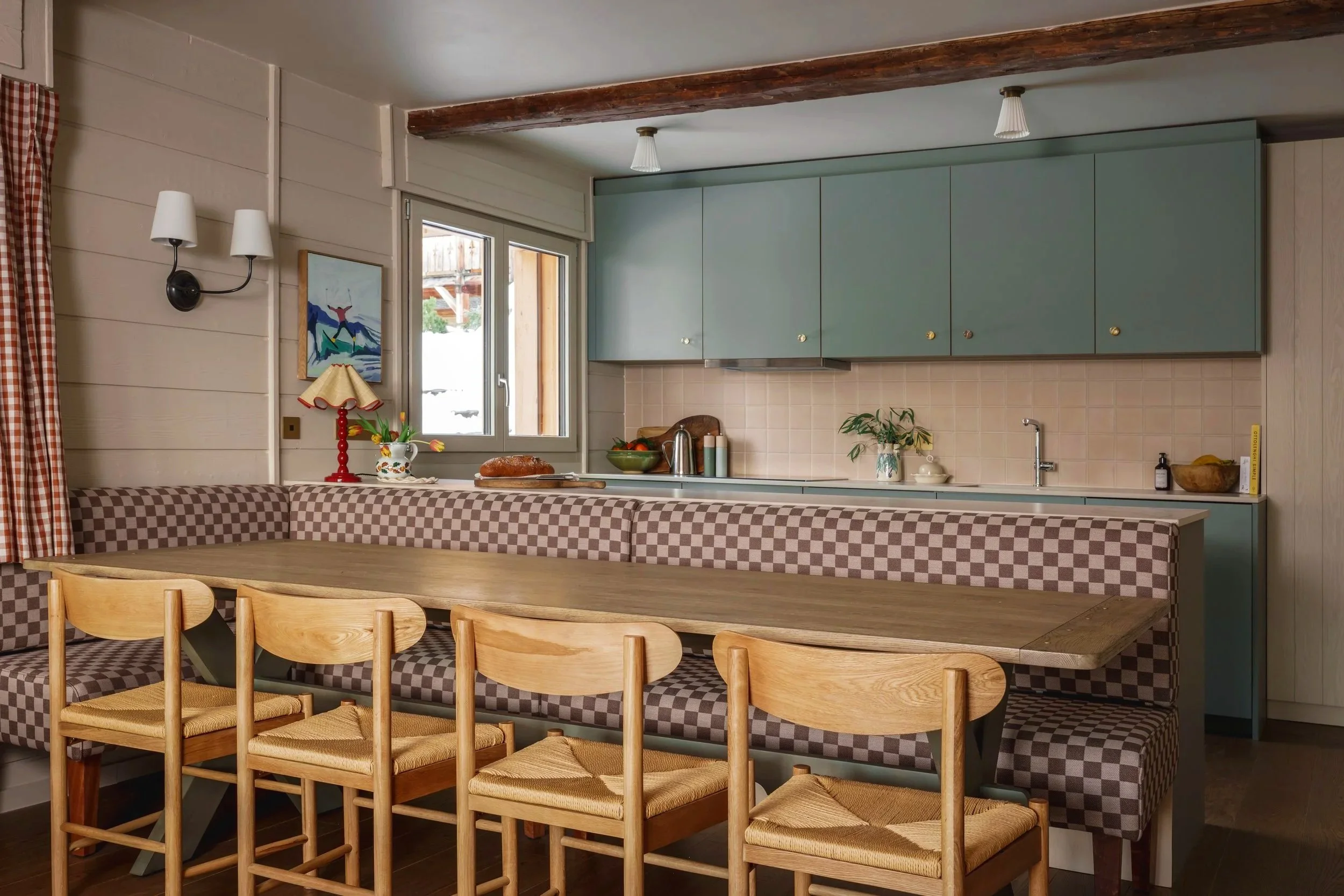 Kitchen with checkered upholstered seating, wooden dining table with four chairs, light blue cabinets, beige tile backsplash, window, and decorative items.