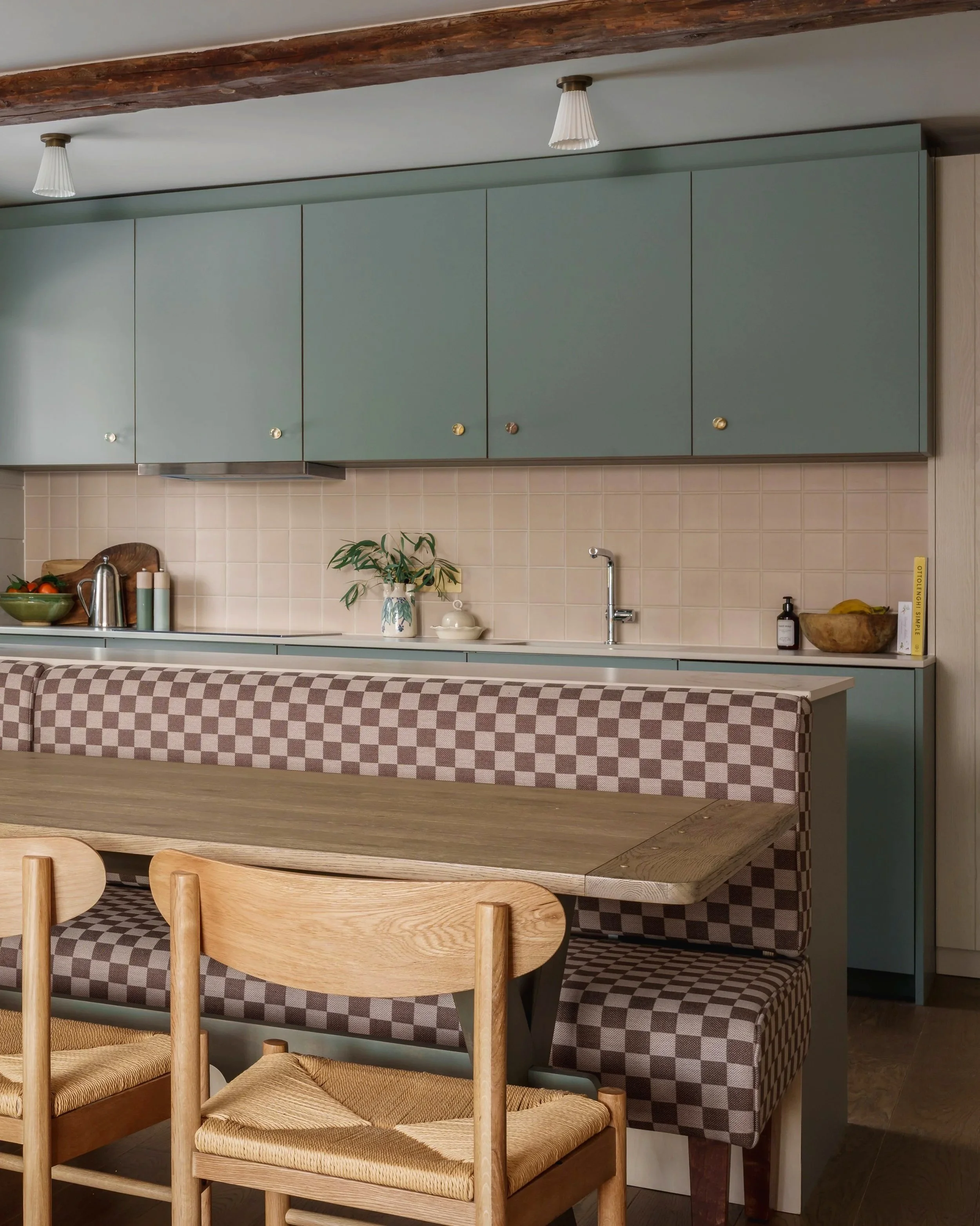 A kitchen with green upper cabinets, a beige tiled backsplash, a wooden dining table, and woven chairs with checkered upholstery.