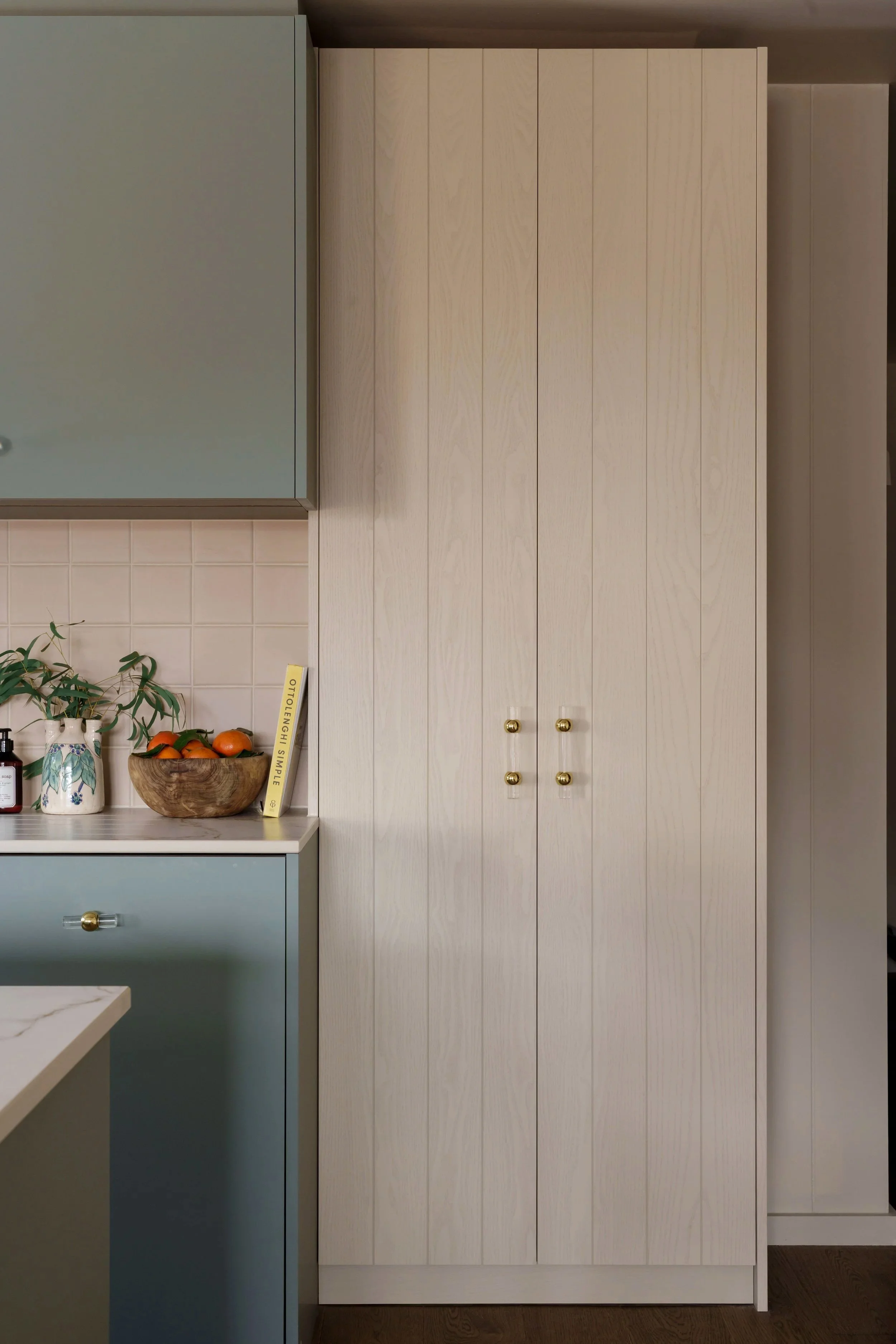 A tall, white wooden cabinet with four vertical panels and round gold knobs, positioned next to a blue cabinet in a kitchen.