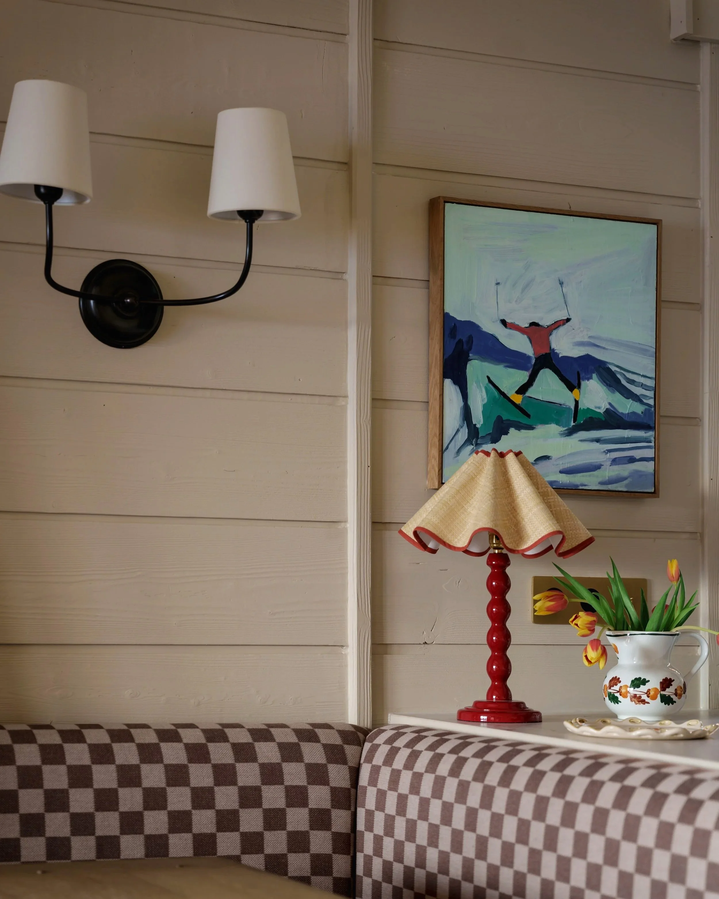 Interior corner of a room with beige wood-paneled walls, a black wall sconce with two white shades, a table lamp with a red base and beige shade, a framed painting of a skier on mountains, a vase with yellow and red tulips, and a checkered brown and 