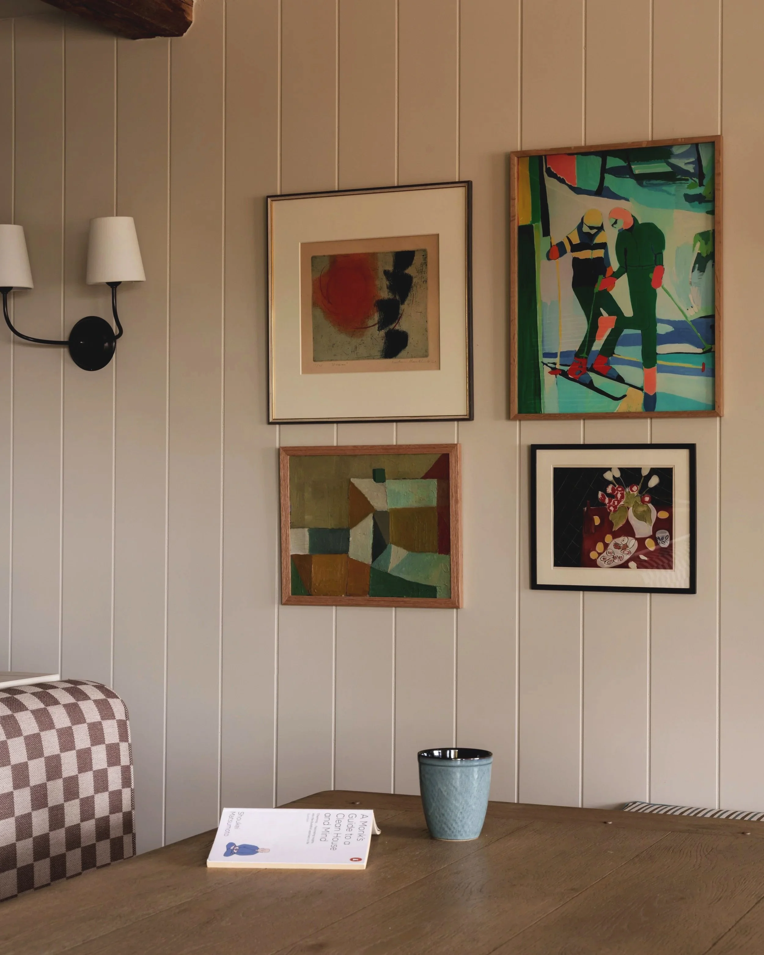 A wall with four framed pieces of art in various styles, a wall-mounted sconce with two white lamps, a wooden table with a book and a blue mug, and a checkered upholstered chair.