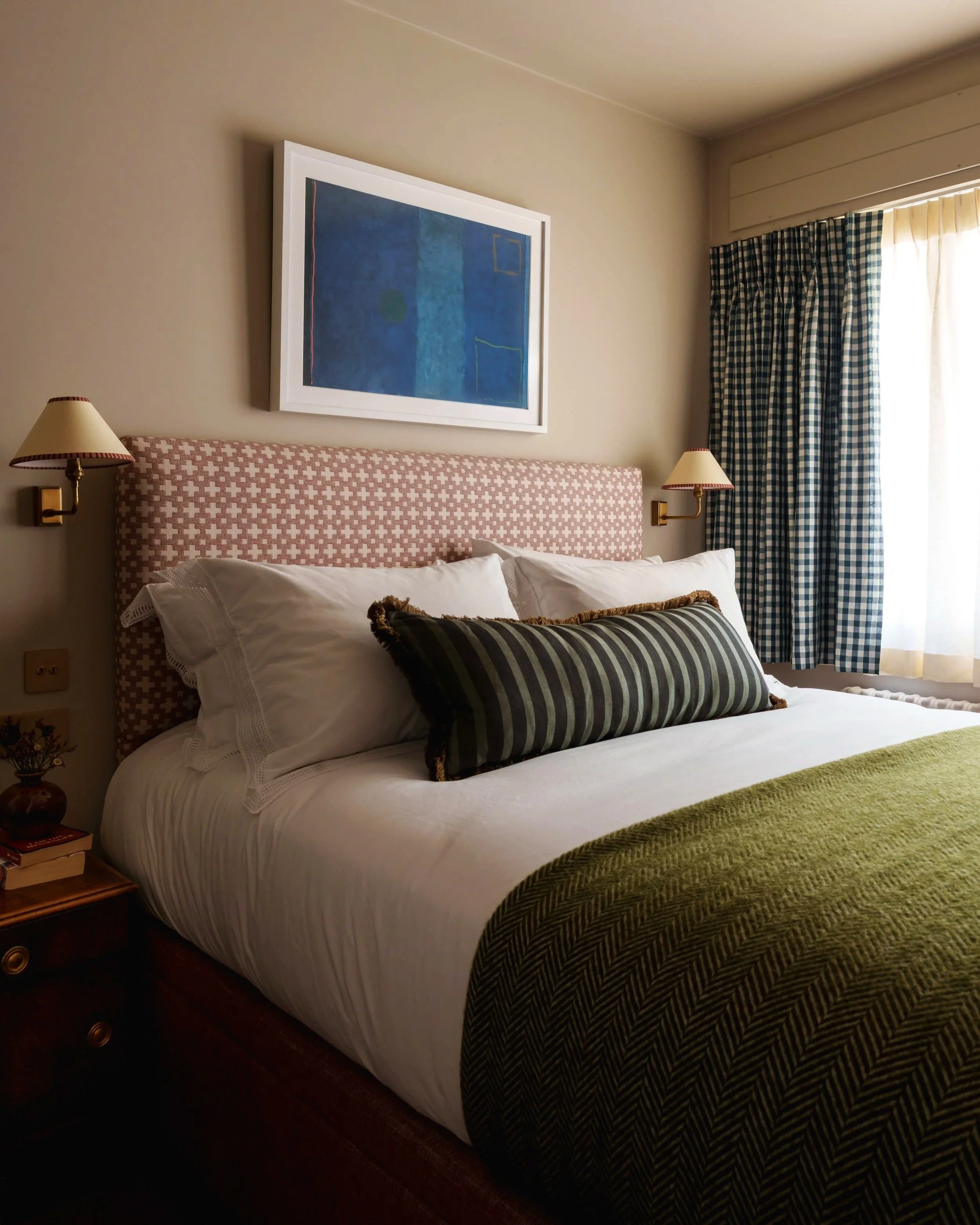 A cozy bedroom with a bed, pink patterned headboard, white pillows, and a long black and green striped decorative pillow. There are two wall-mounted lamps on either side of the bed, a framed abstract blue painting above, and a window with blue and wh