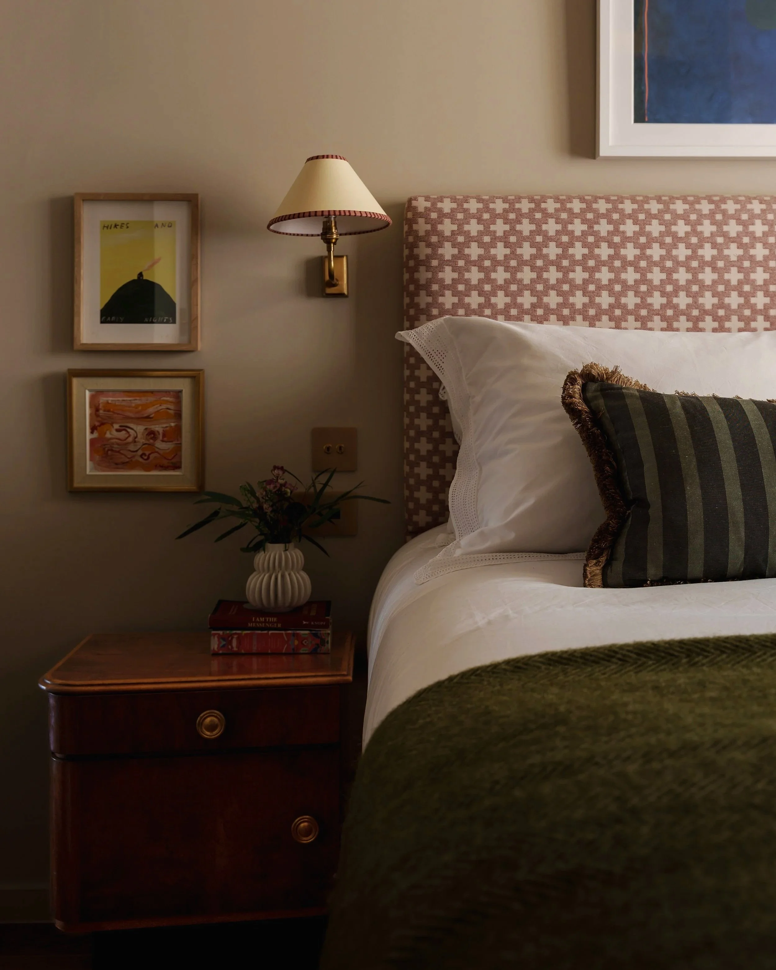 A cozy bedroom corner featuring a bedside table with a flower vase and books, a bed with patterned pillows, and wall art with a wall-mounted lamp.