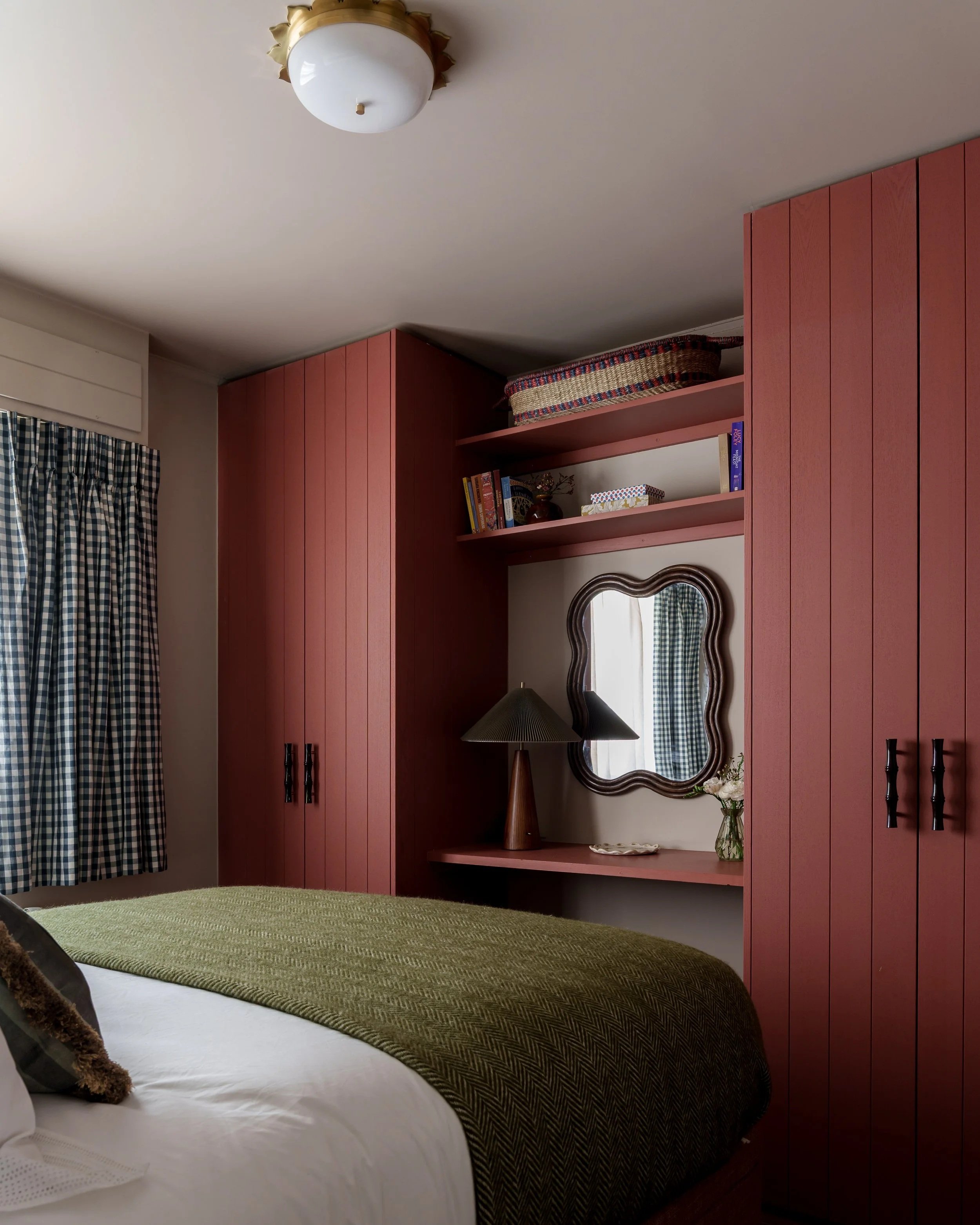 A cozy bedroom with a bed covered in a green blanket, a wooden bedside table, a lamp, a small mirror, and built-in red wooden cabinets. There are patterned curtains on the window.