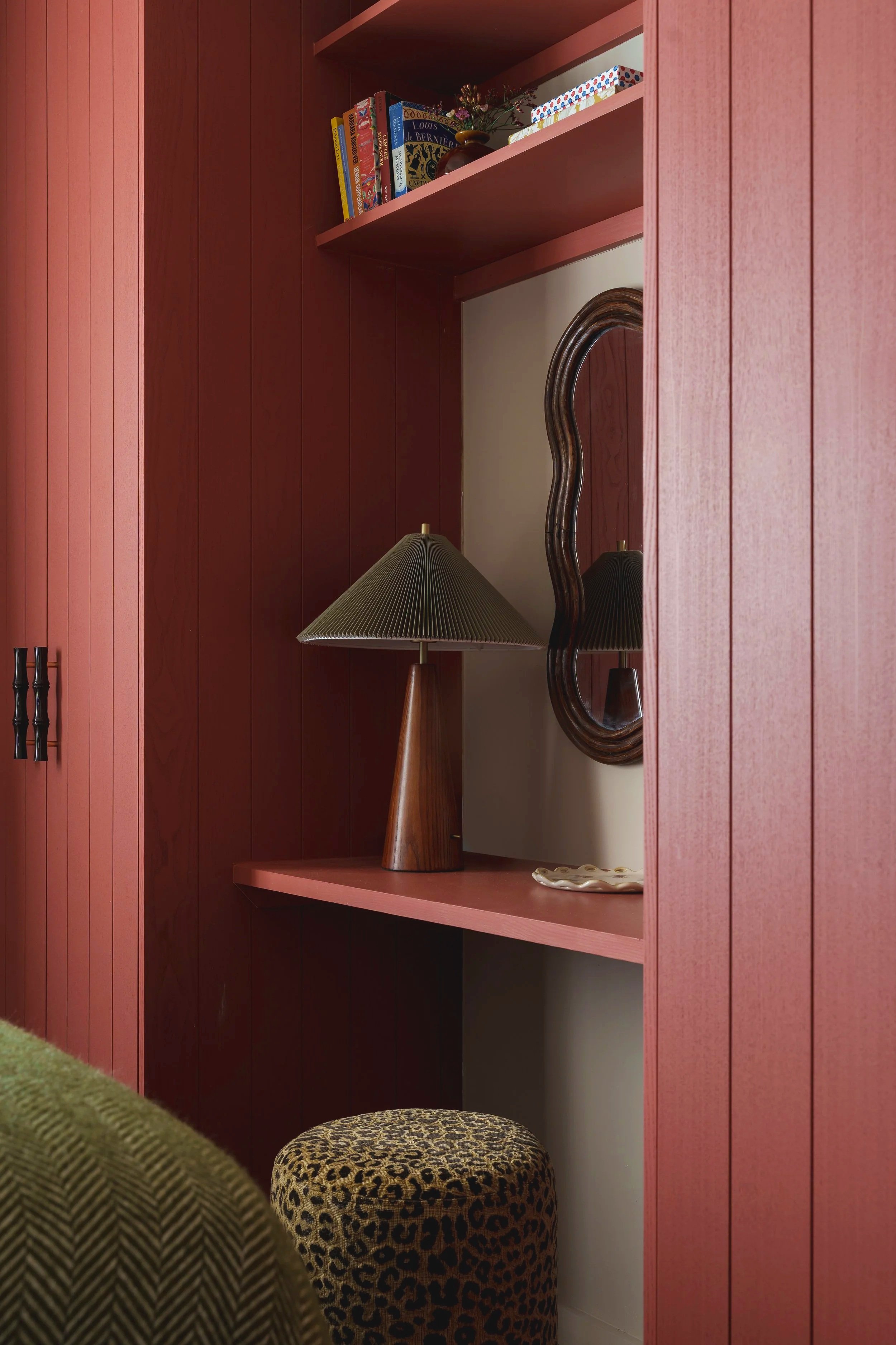 Interior corner with a pink wooden wall and built-in shelves. The top shelf holds books and a small vase, and the bottom shelf has a vintage lamp with a cone-shaped shade, a decorative dish, and a wavy mirror. A leopard print ottoman is below the low