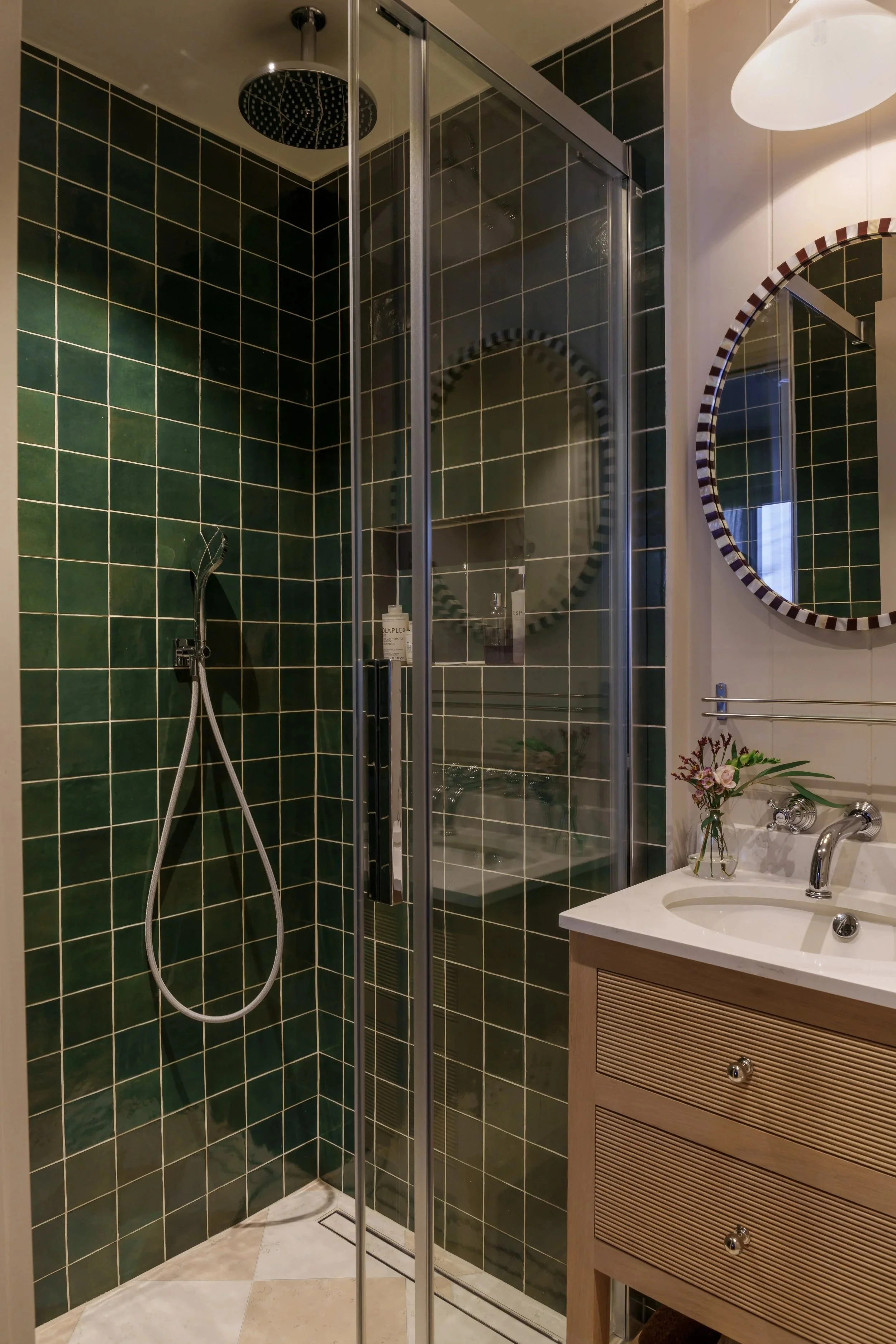 A bathroom with green tiled walls, a glass shower with a rain showerhead, a white sink with a wooden cabinet, a mirror, and a vase with flowers on the countertop.