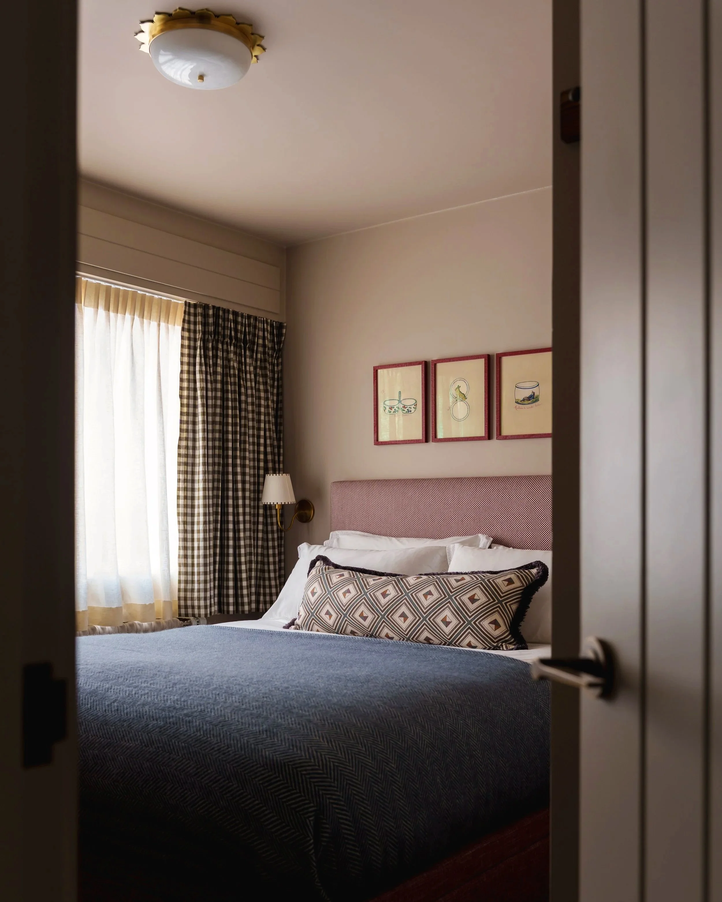 A bedroom with a bed, patterned pillows, framed art on the wall, a window with curtains, and a ceiling light.