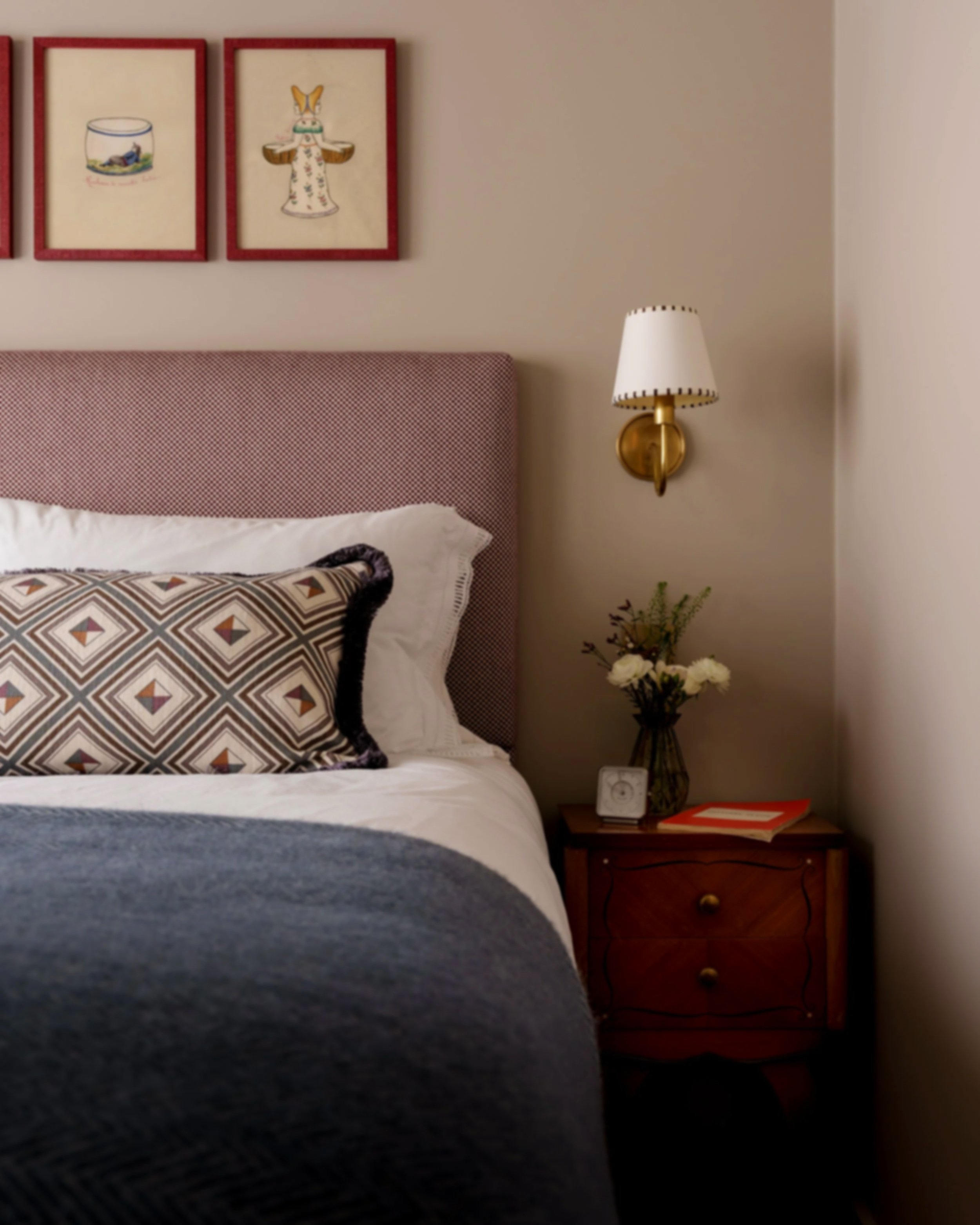 A cozy bedroom corner with a bed that has white sheets, a patterned pillow, and a pink upholstered headboard. A wooden nightstand with a small vase of white flowers, a clock, and a red book. A wall-mounted lamp with a white shade and brass arm. Three
