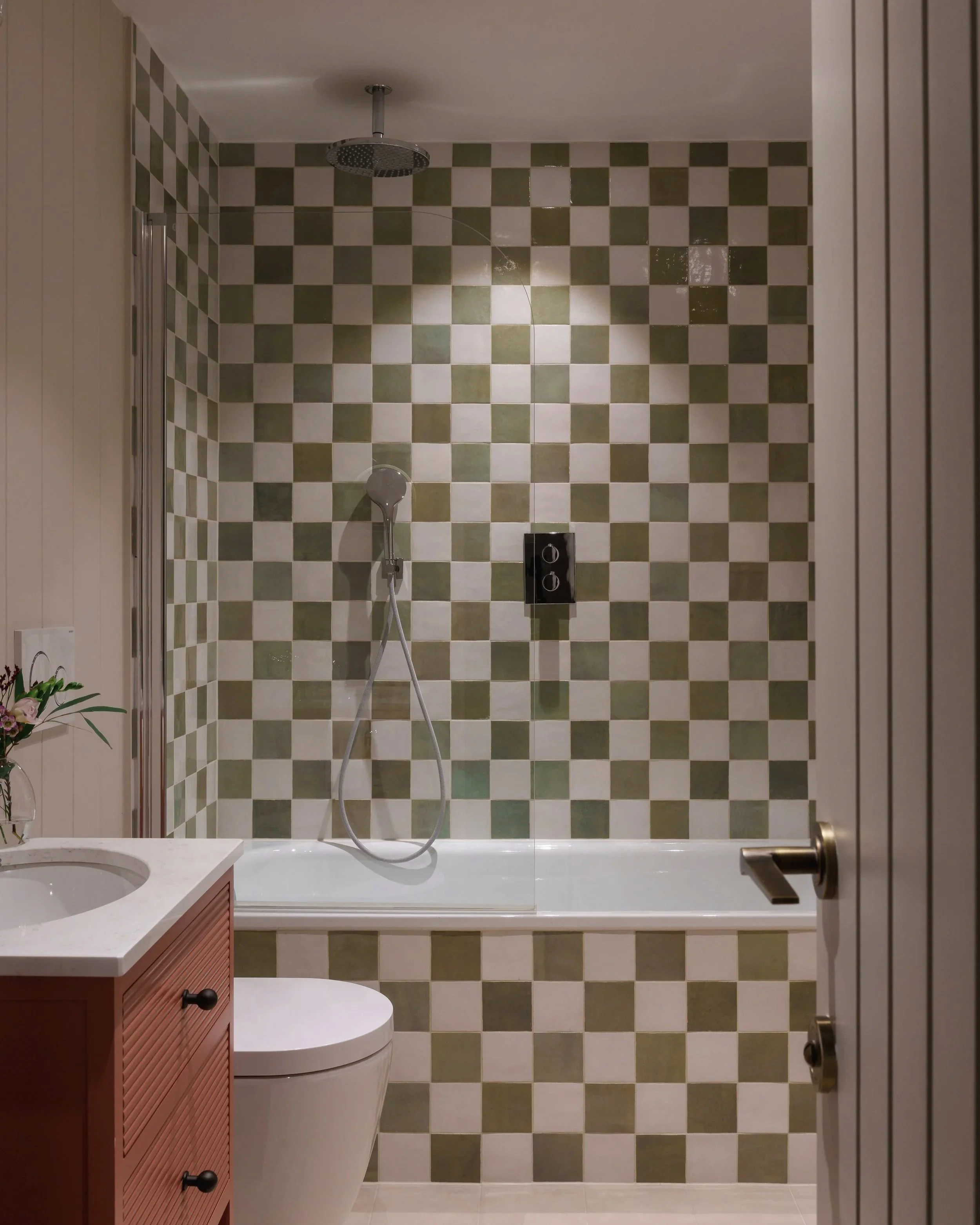 Bathroom with checkered green and white tiled shower wall, showerhead, handheld shower, black control panel, white bathtub, toilet, pink vanity with white countertop, flower arrangement, and partially open white door.